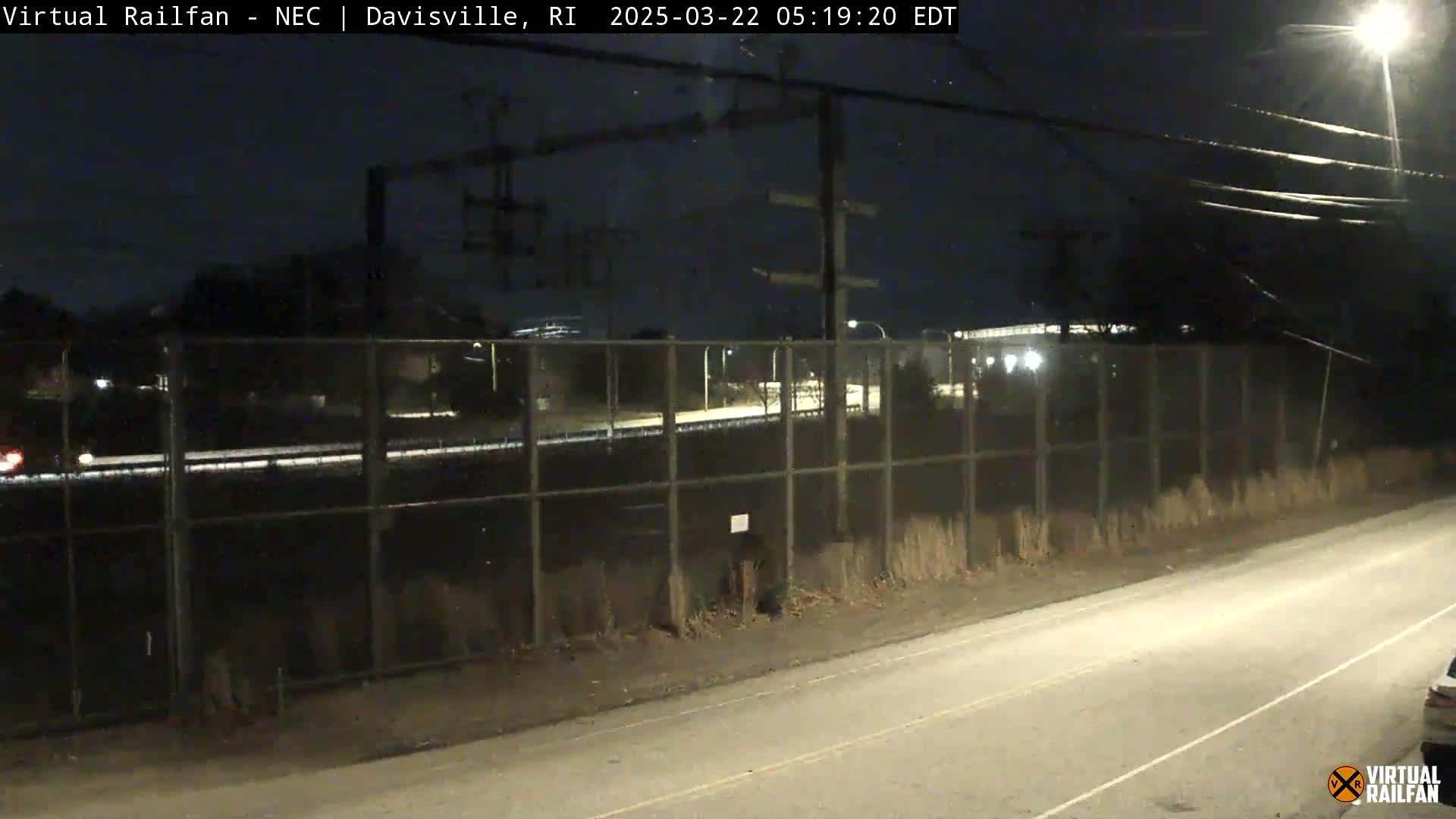 A nighttime view of a highway and a chain-link fence separating it from a dark, industrial area under a clear, dark sky.