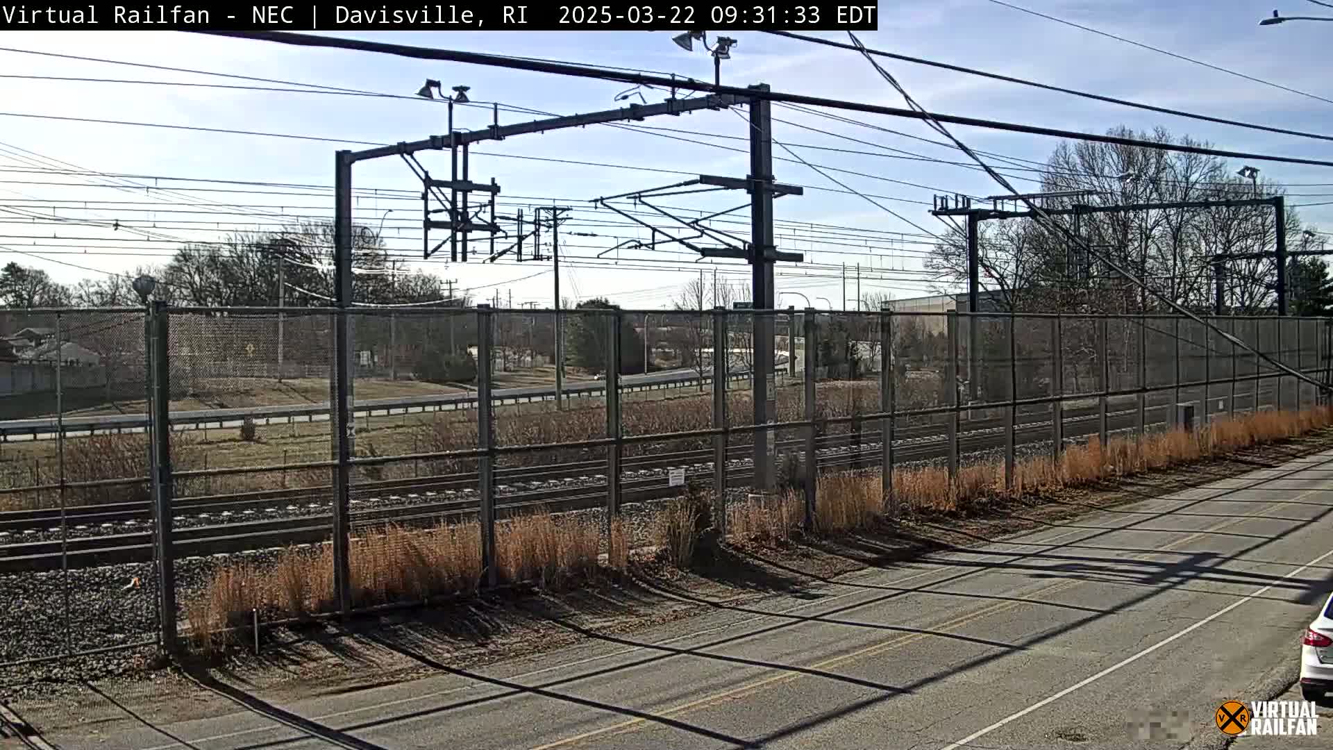 A view of a railway line behind a chain-link fence on a sunny day, with a road and sparse vegetation visible in the foreground.