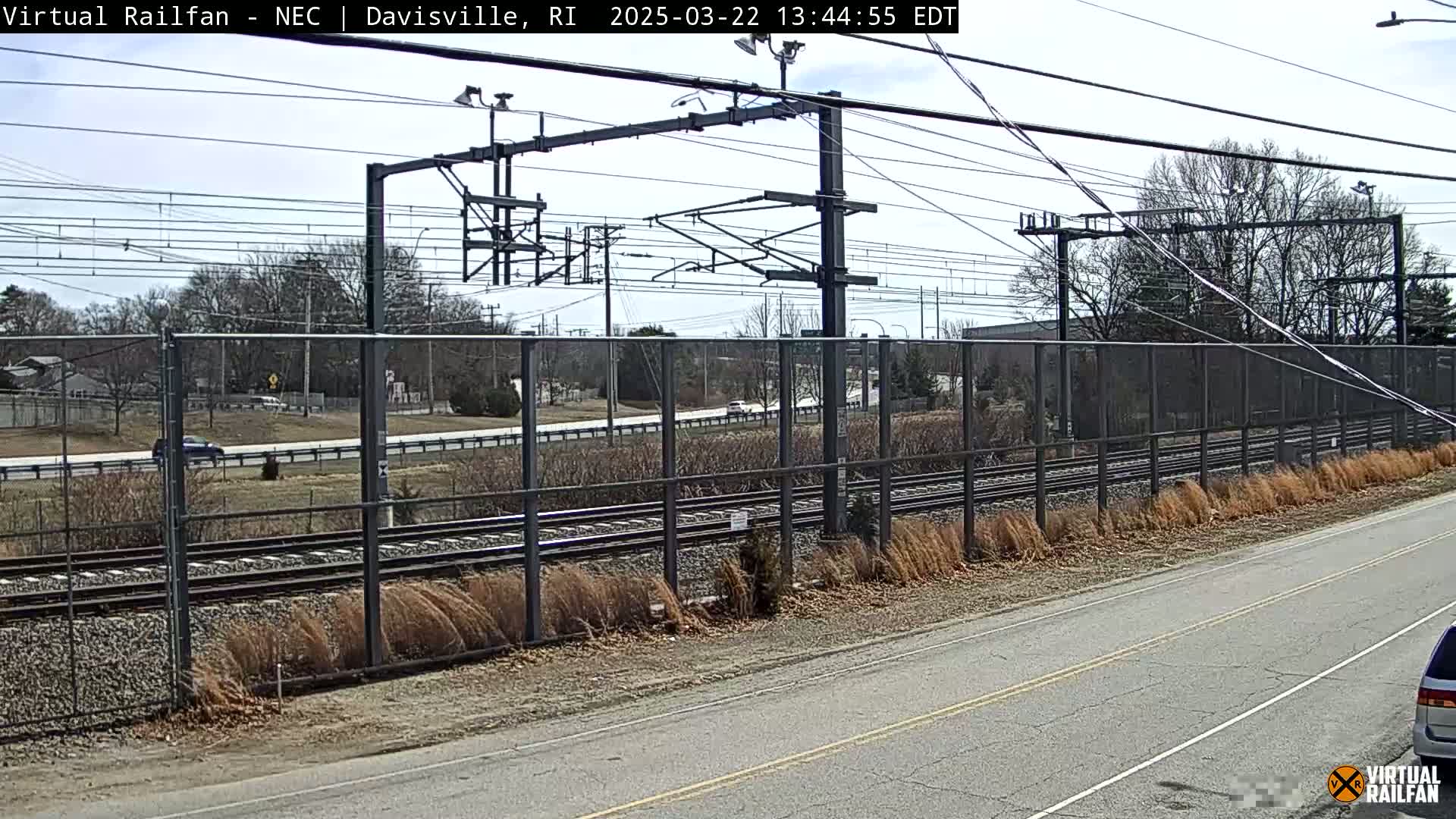 A clear, sunny day reveals a view of train tracks bordered by a chain-link fence, with a road and sparse, wintery vegetation alongside.