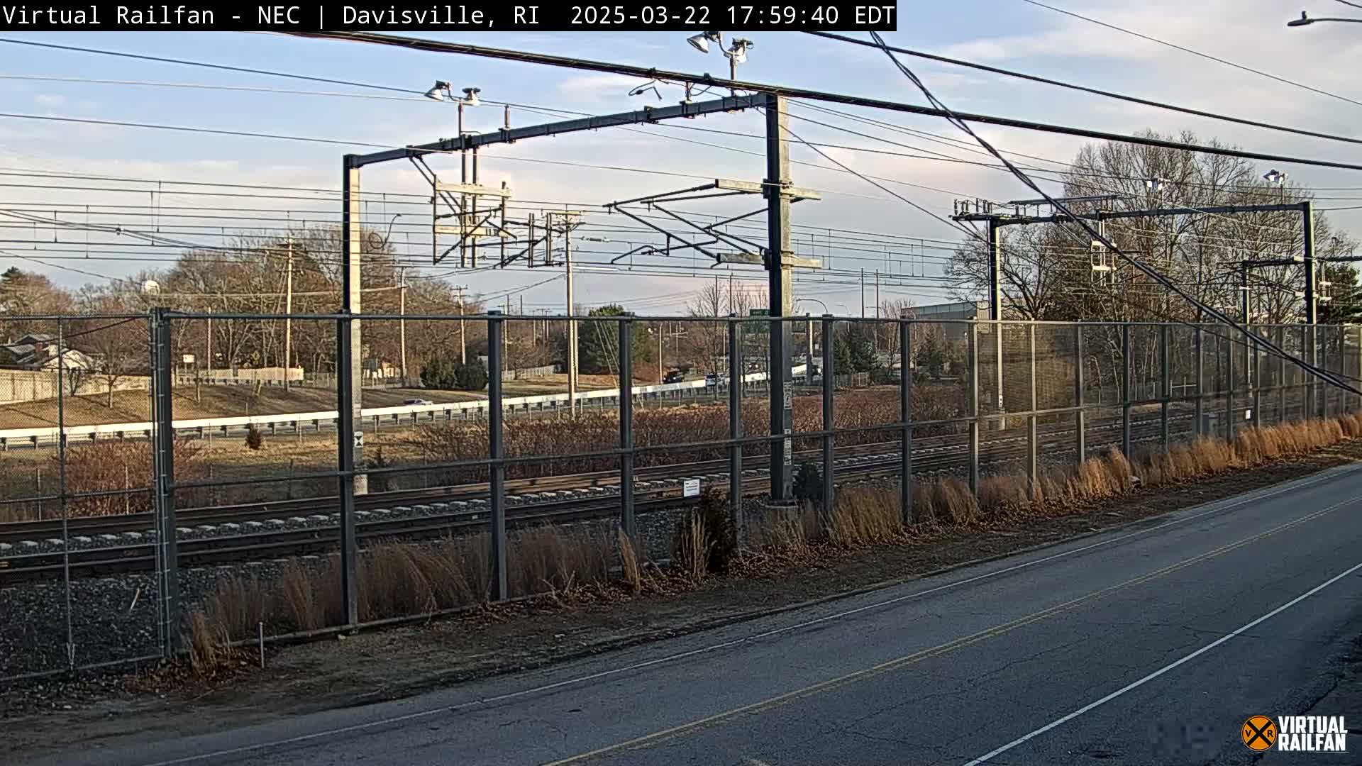 A view of railroad tracks behind a chain link fence, with a road in the foreground, under a mostly clear sky.