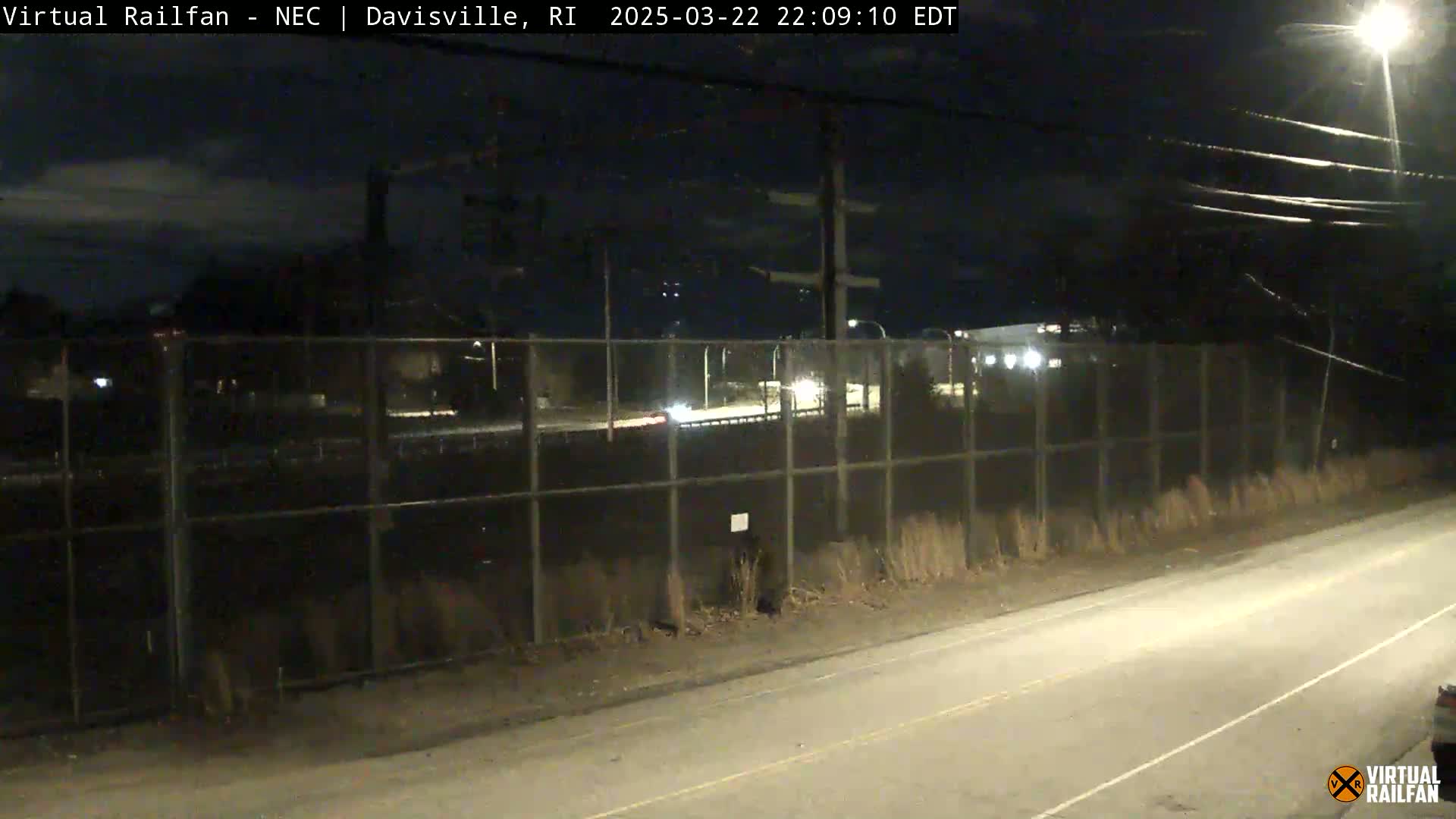 A nighttime view of a road next to a chain-link fence, with a few vehicles visible in the distance under a partly cloudy sky.