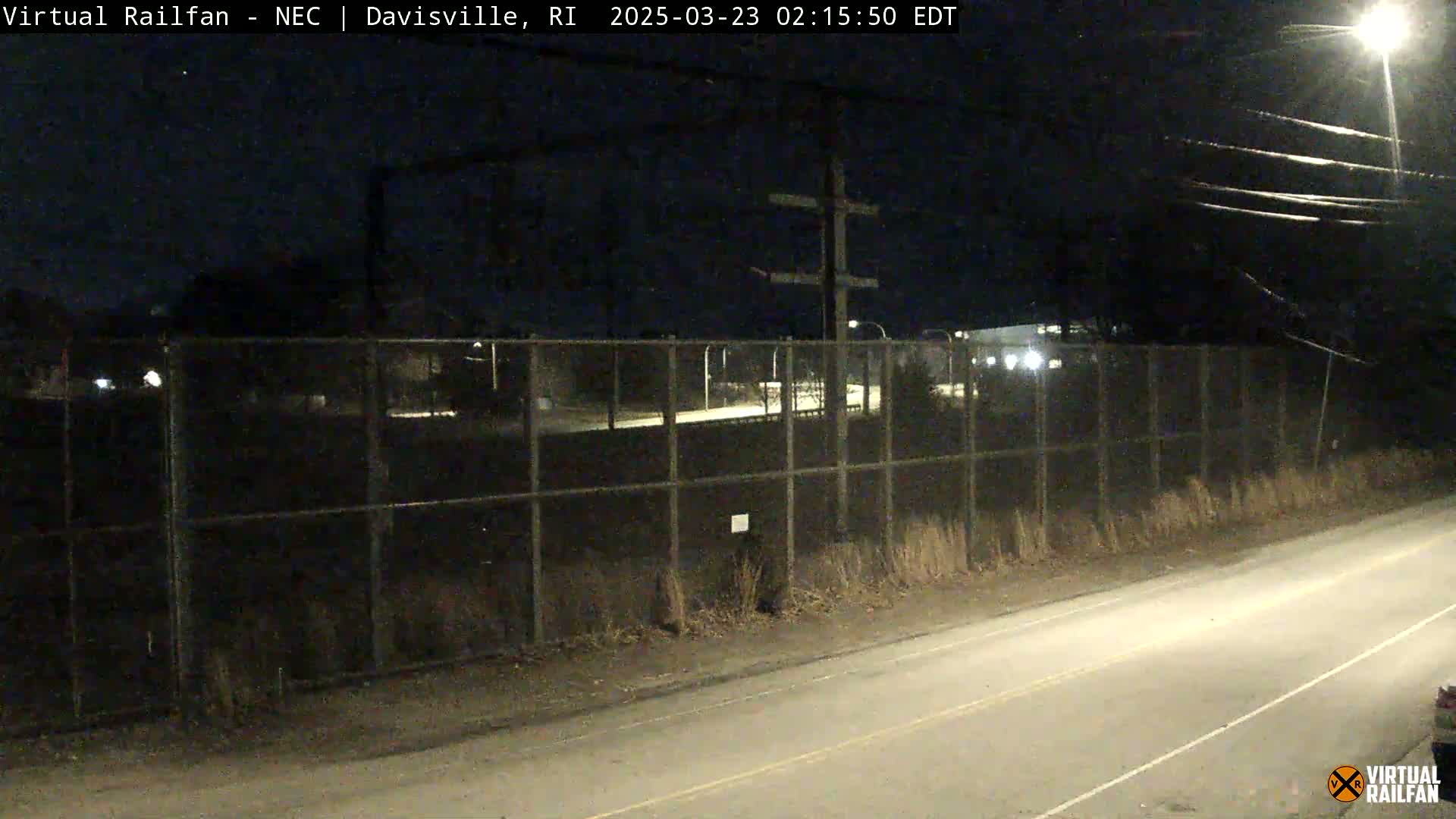 A nighttime view of a chain-link fence bordering a road, with power lines and dimly lit buildings beyond the fence.