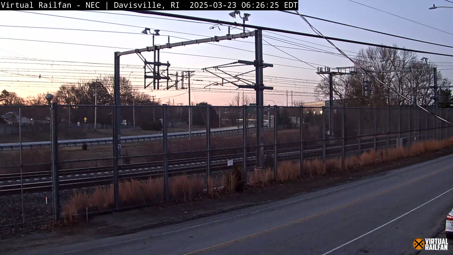 A twilight view of railroad tracks behind a chain link fence, next to a road, with bare winter trees in the background.