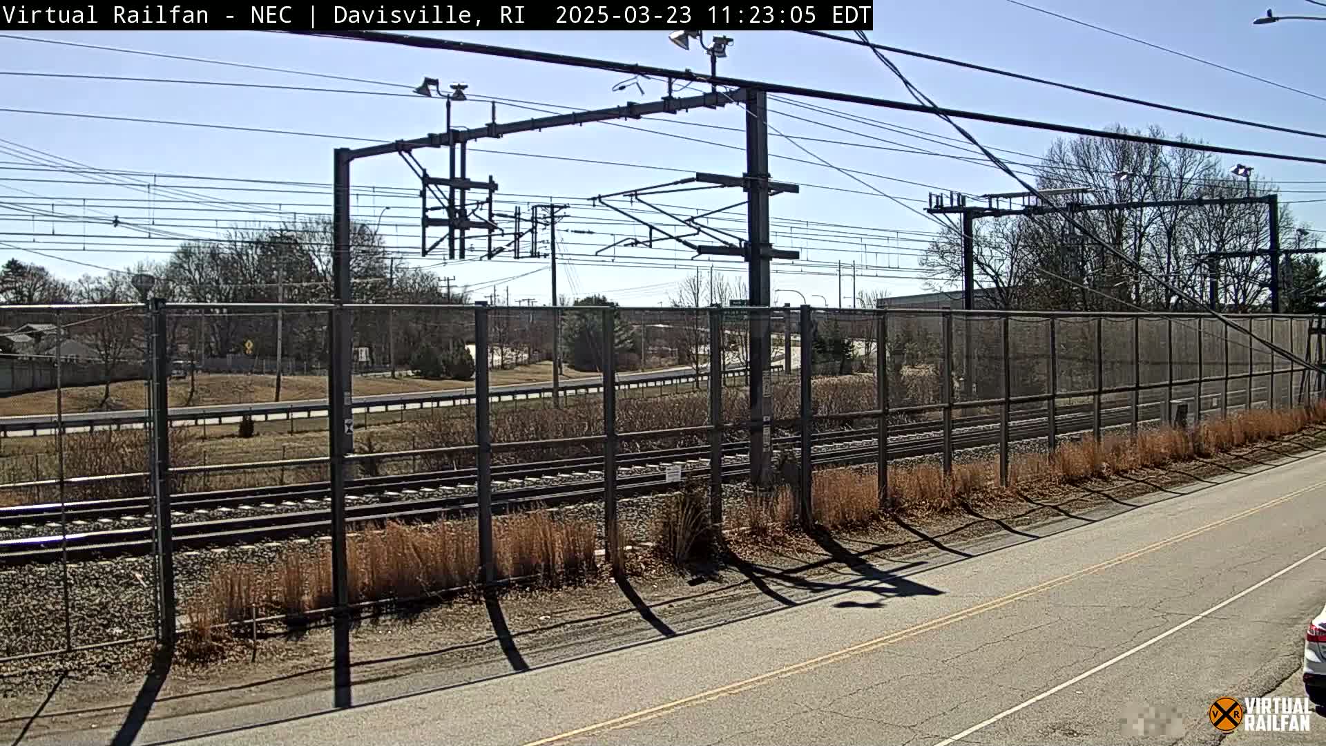 A sunny view of railroad tracks behind a chain link fence, next to a road, with trees and buildings in the distance.