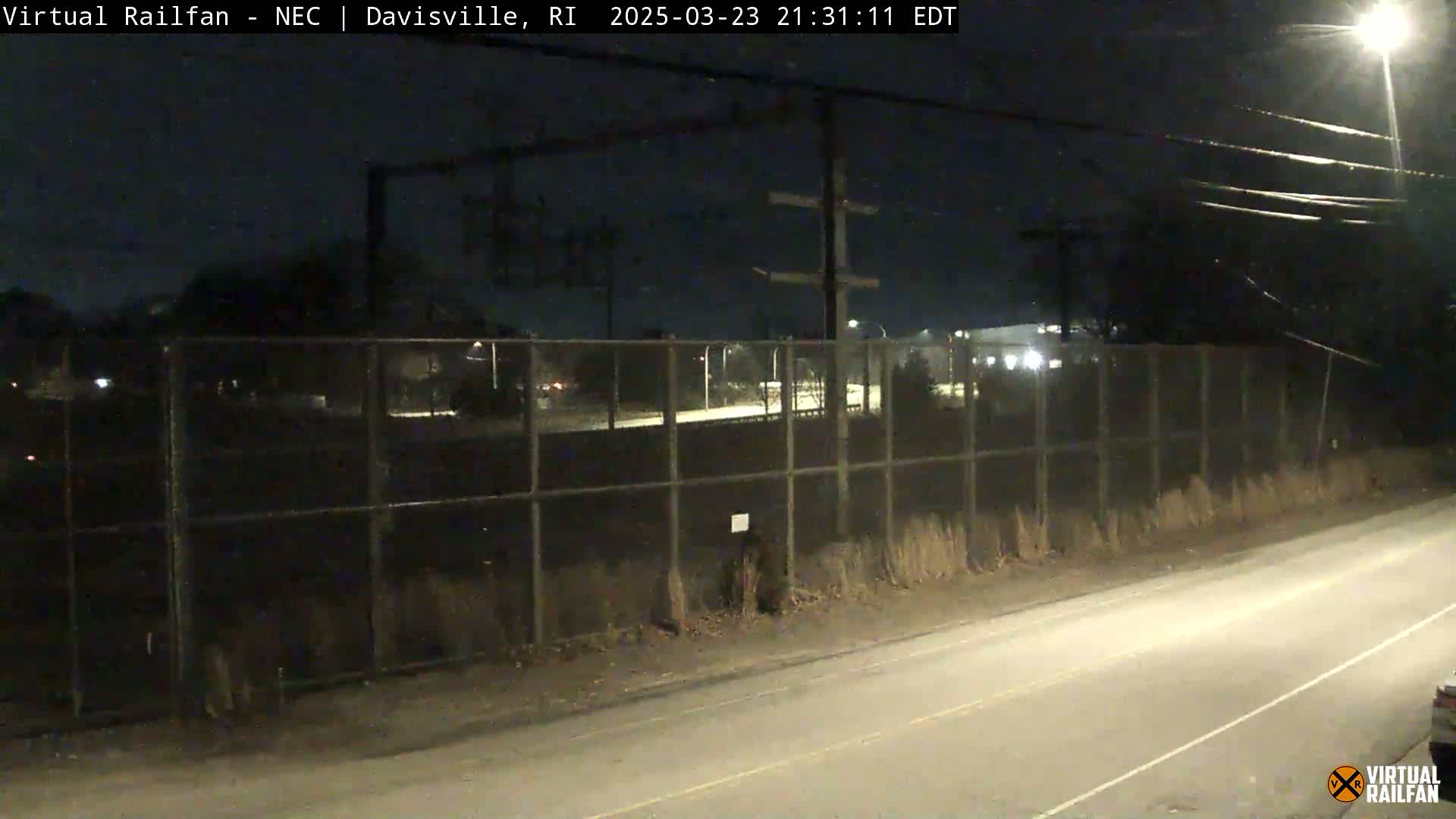A nighttime view shows a road next to a tall chain-link fence surrounding a dark area, with streetlights illuminating the scene under a clear, dark sky.