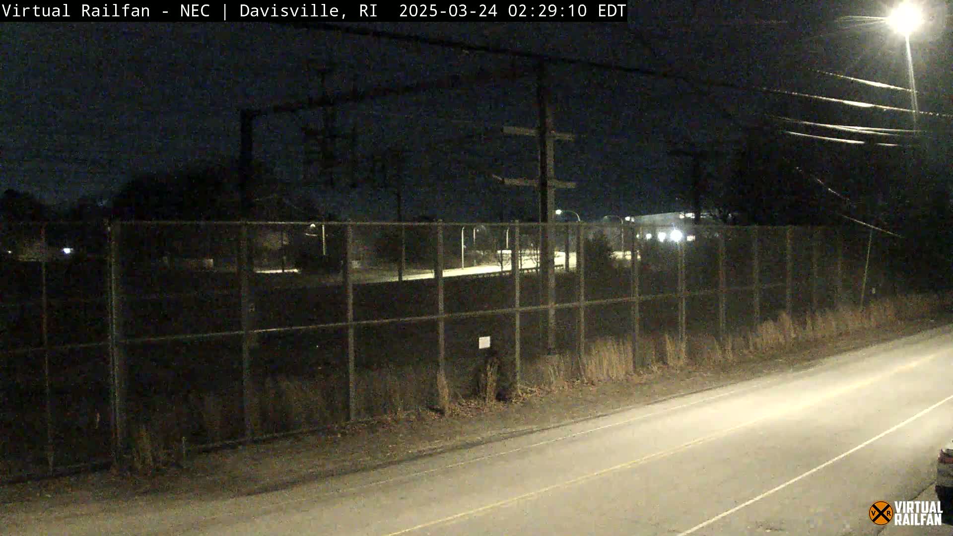 A nighttime view of a chain-link fence enclosing a dark area beyond a road, with power lines overhead and a streetlight illuminating part of the scene.