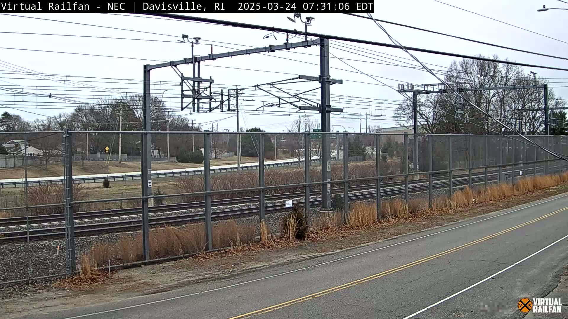 A view of railroad tracks behind a chain link fence next to a road on an overcast day.
