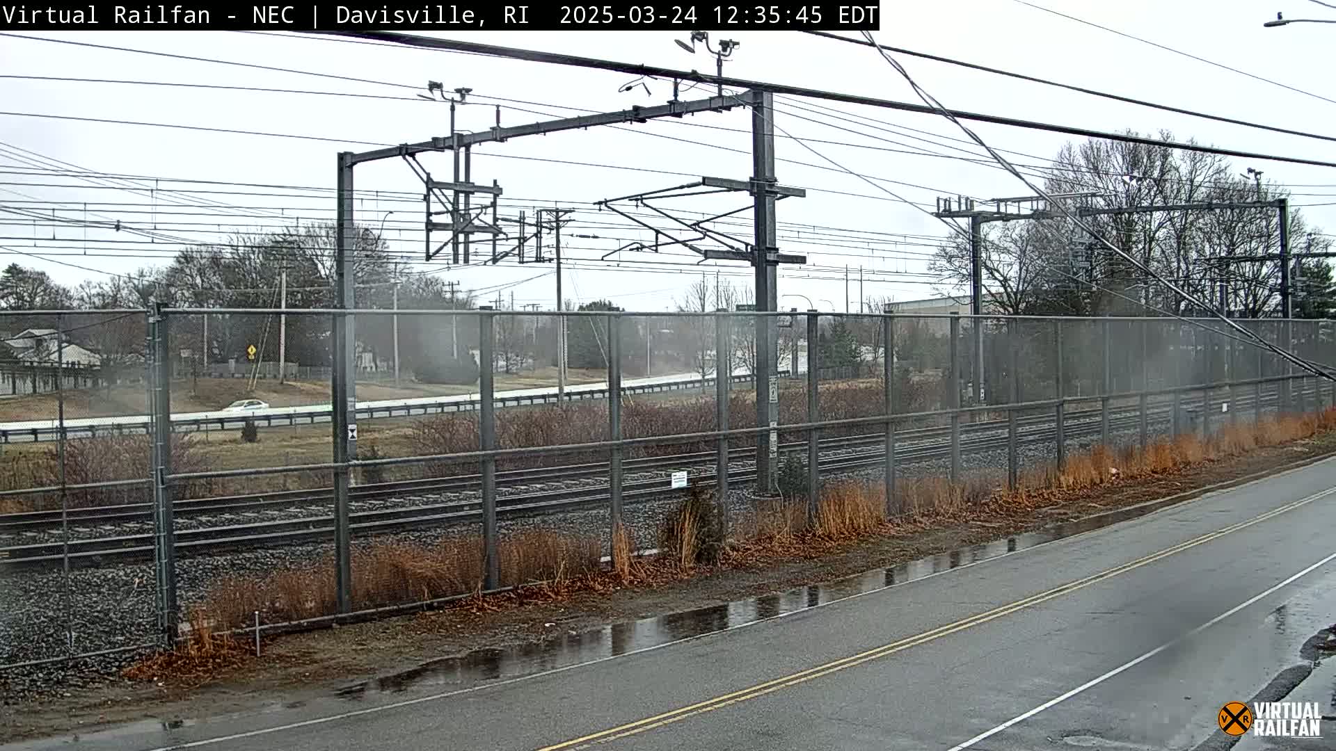 A wet road runs beside railroad tracks fenced off from a roadway beyond, on an overcast day.