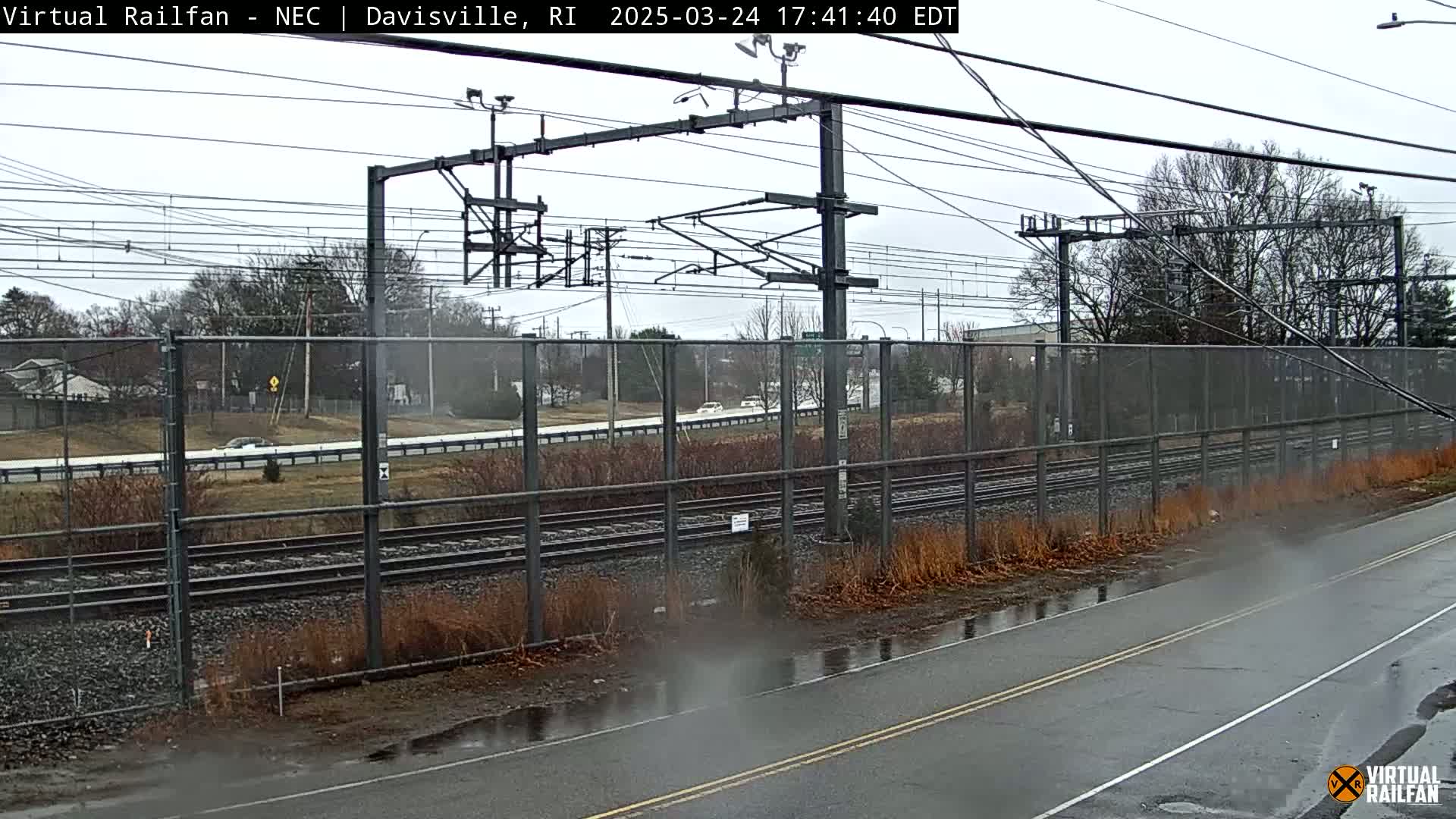 A wet road next to train tracks runs alongside a chain-link fence separating it from a roadway with cars, under an overcast sky.