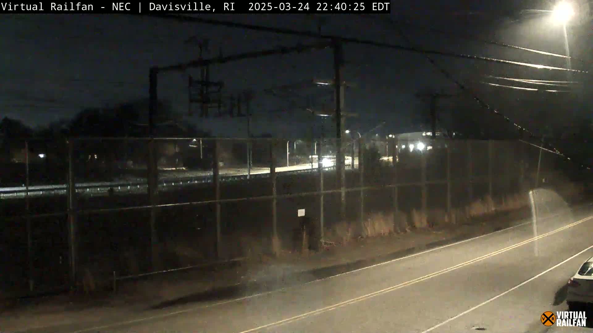 A nighttime view of a roadway next to a chain-link fence, with a vehicle visible in the distance and light rain falling.