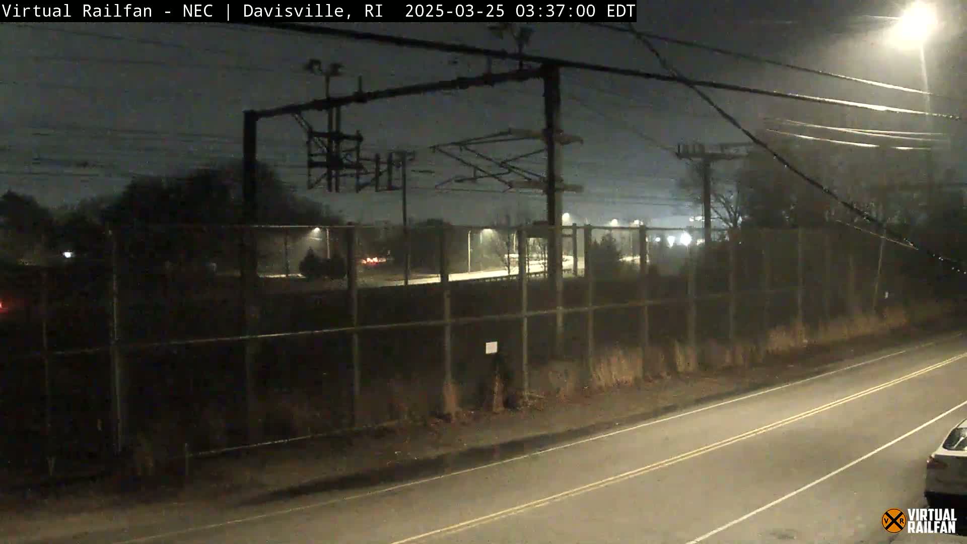 A nighttime view of a rain-slicked road next to a chain-link fence bordering a railway, with overhead power lines and distant lights visible.