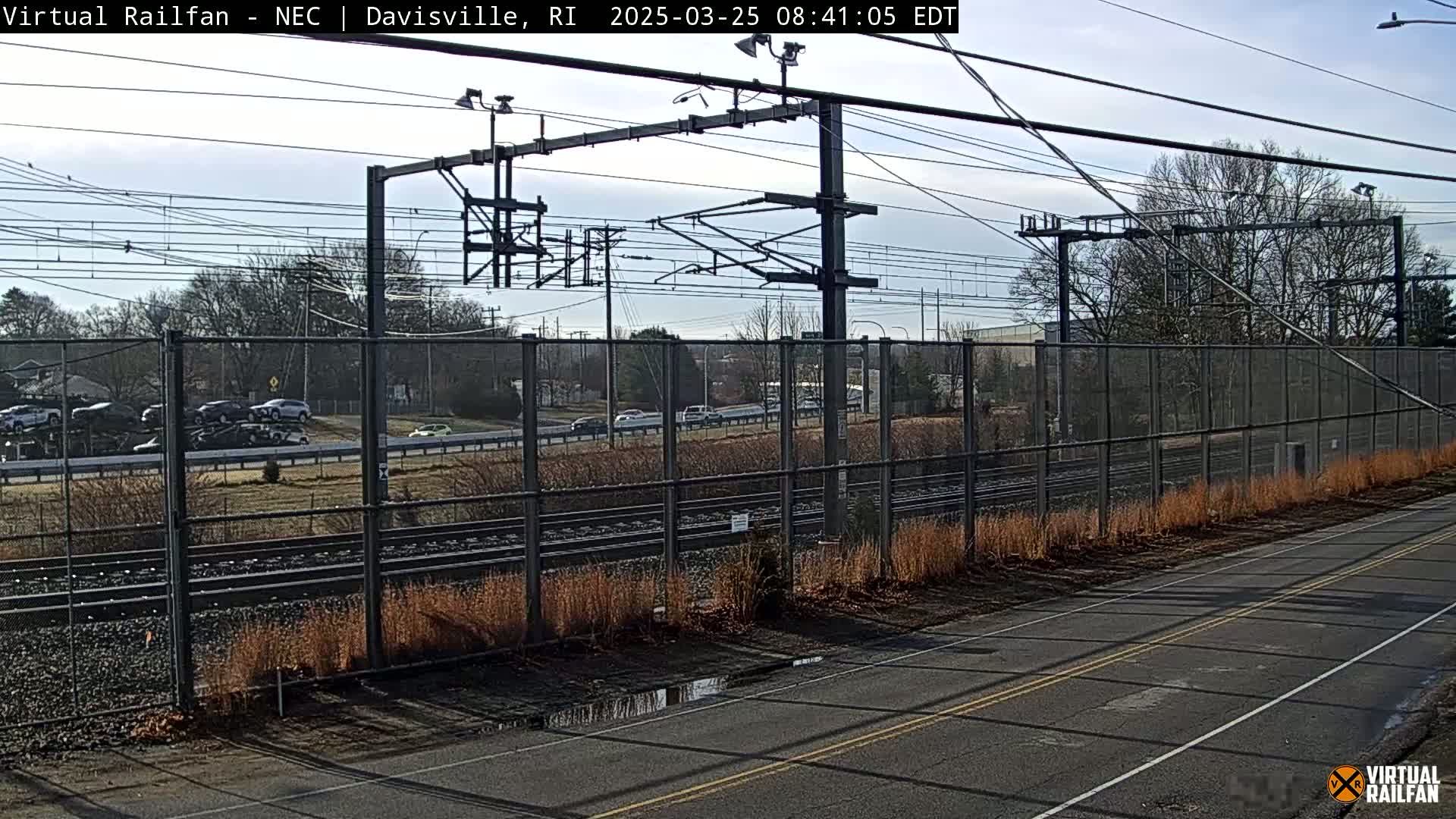 A view of railroad tracks bordered by a chain-link fence and a paved road, under a mostly clear sky with some scattered clouds.