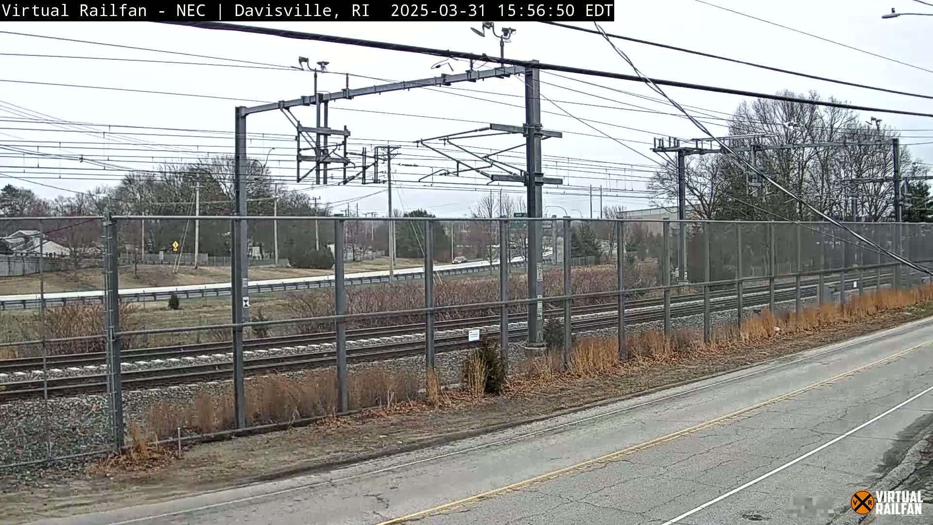 A view of train tracks and a road, separated by a chain-link fence, on an overcast day.