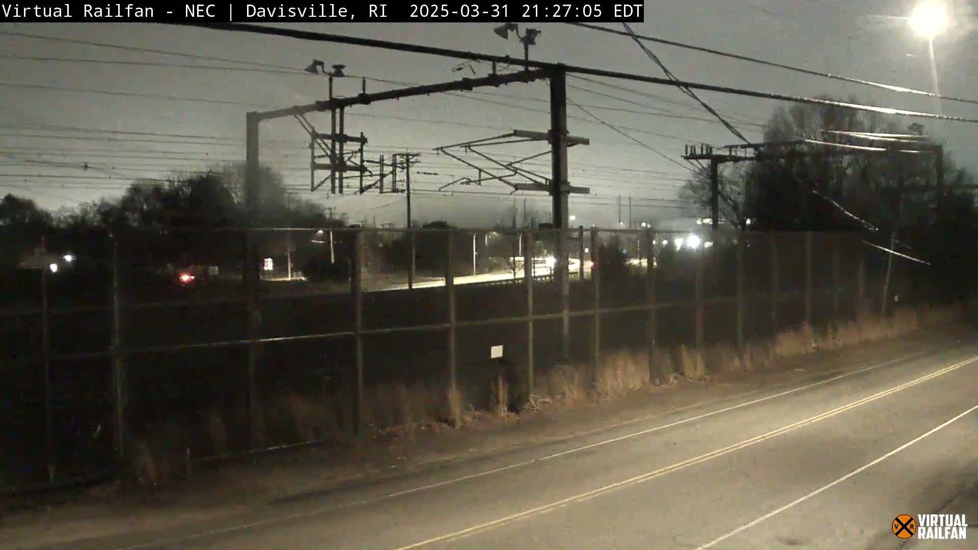 A nighttime view of a railway line behind a chain-link fence, with a road in the foreground under a cloudy sky.