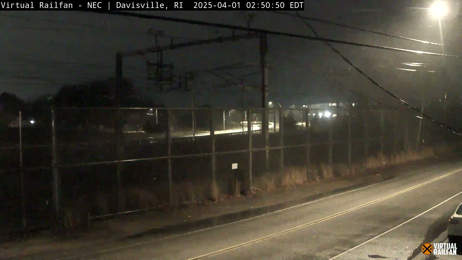 A nighttime view of a rain-slicked road next to a chain-link fence, beyond which a train track and some lights are visible under a dark, overcast sky.
