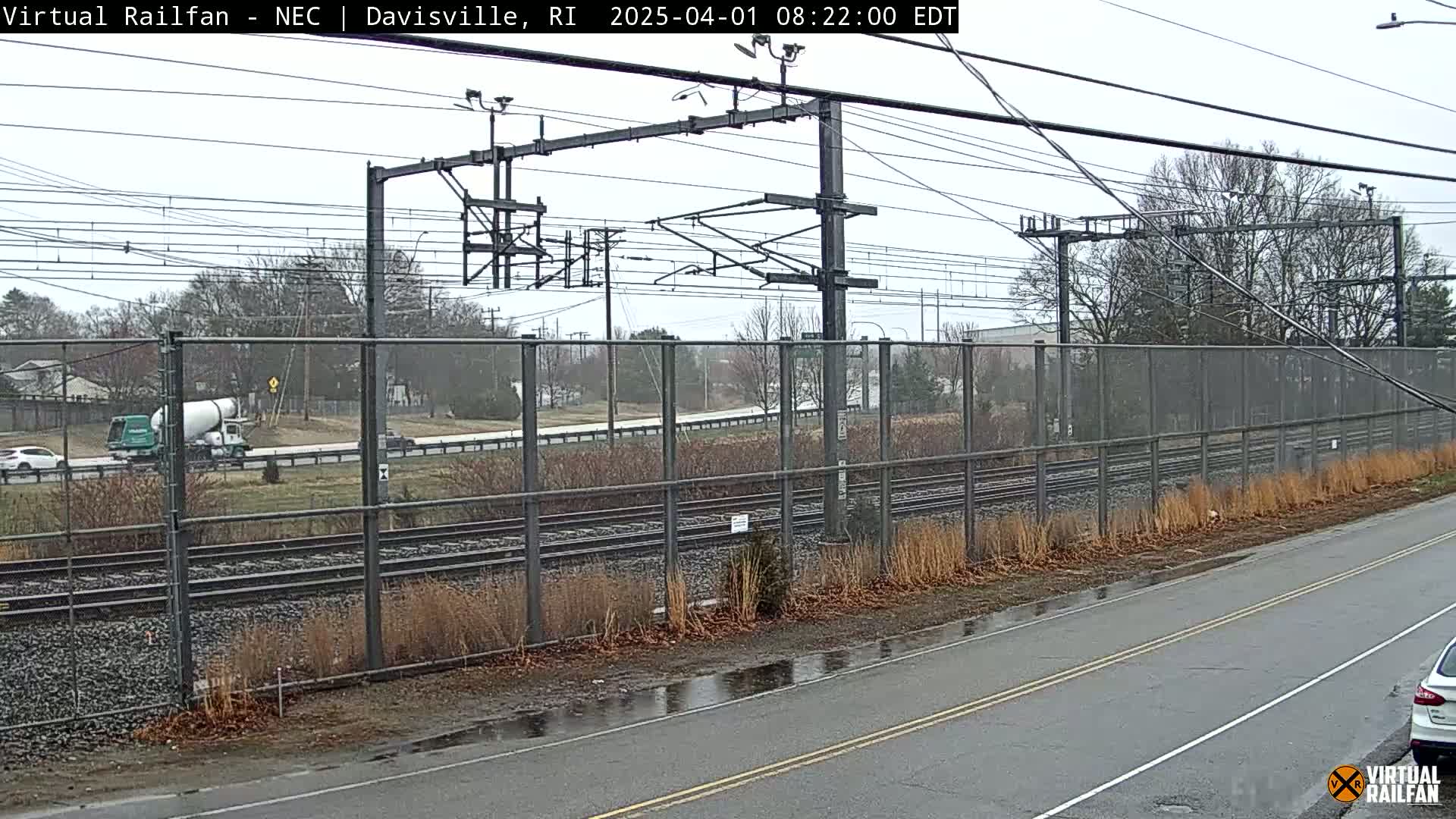 A cement mixer truck drives past a railway line on an overcast day, next to a chain-link fence separating the road from the tracks.