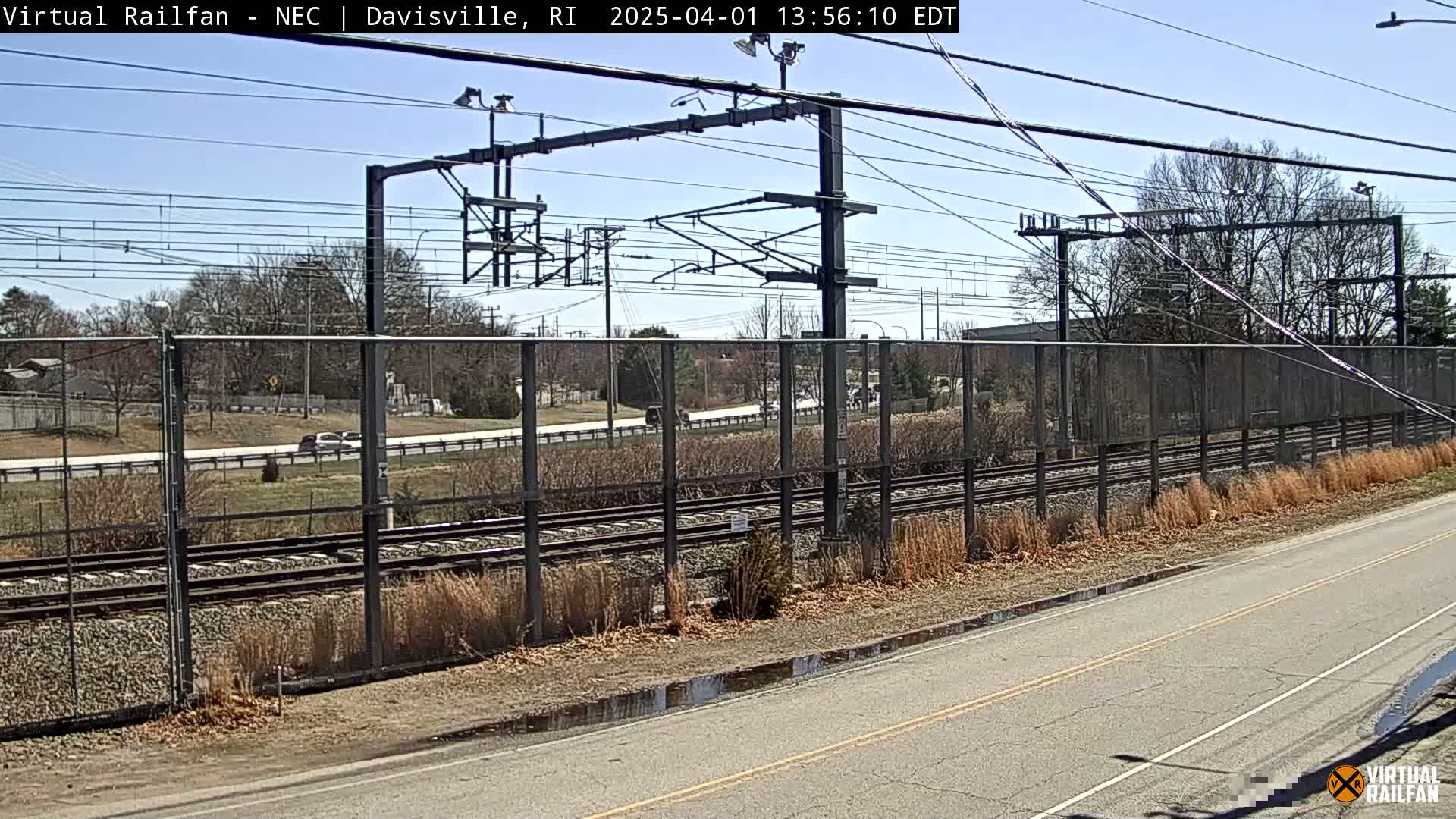 A view of train tracks separated from a road by a fence, under a clear blue sky.