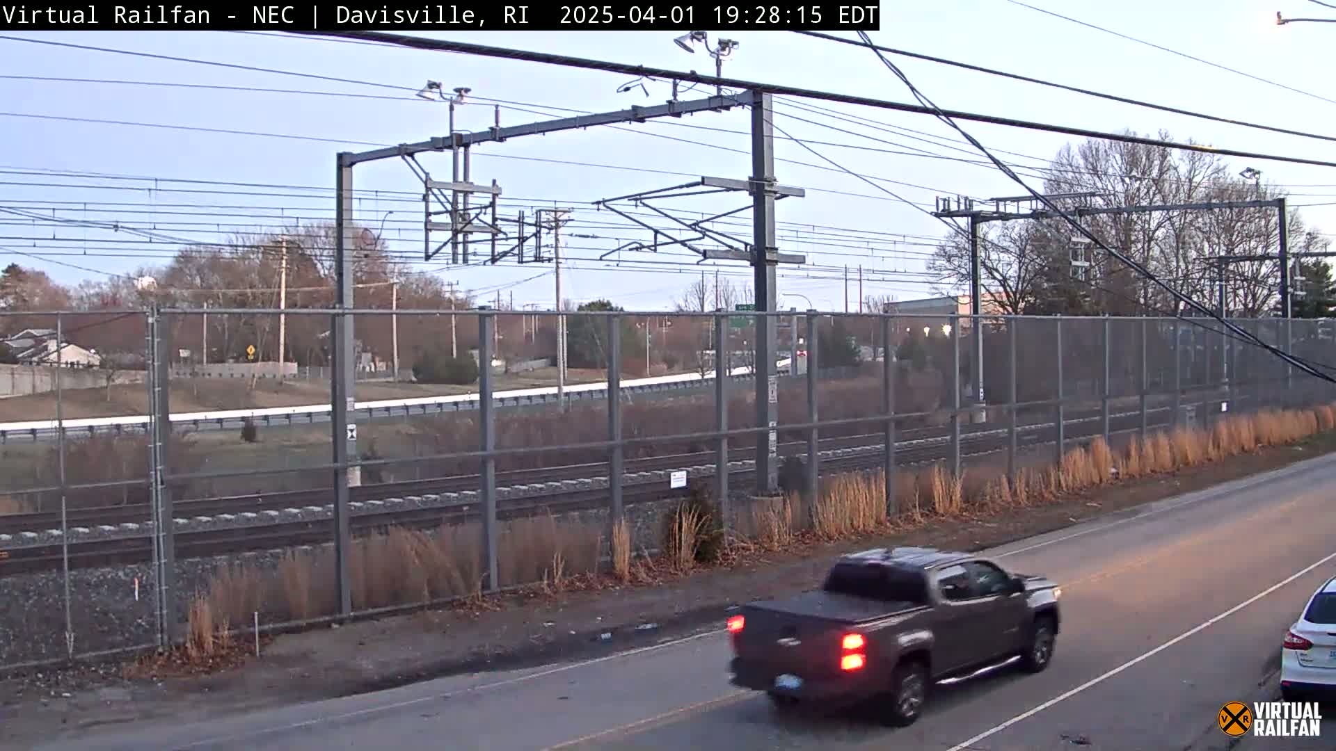 A brown pickup truck drives on a road next to a chain-link fence separating it from a railway line under a clear, twilight sky.