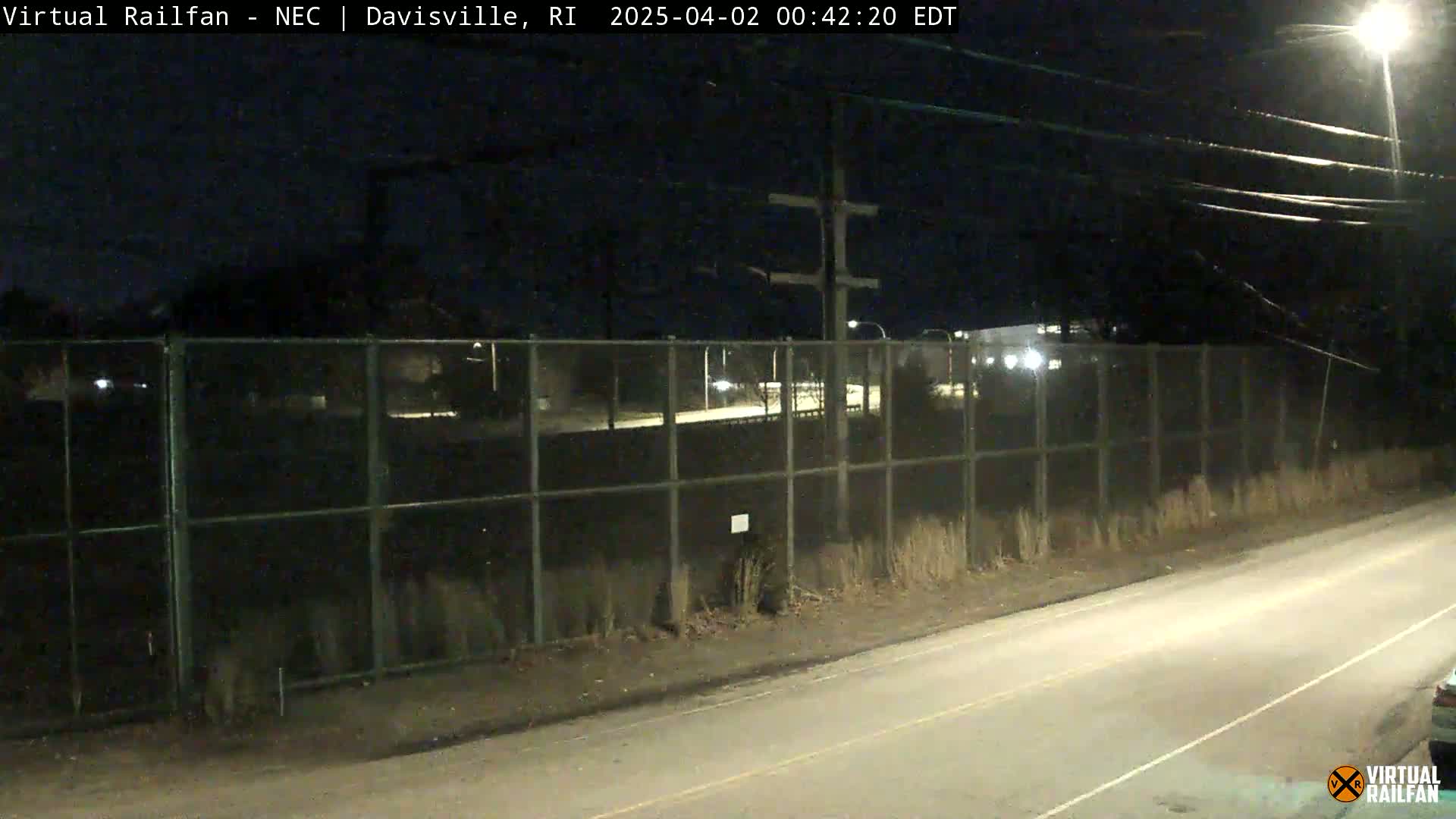 A nighttime view of a chain-link fence separating a road from a rail yard, under a dark sky.