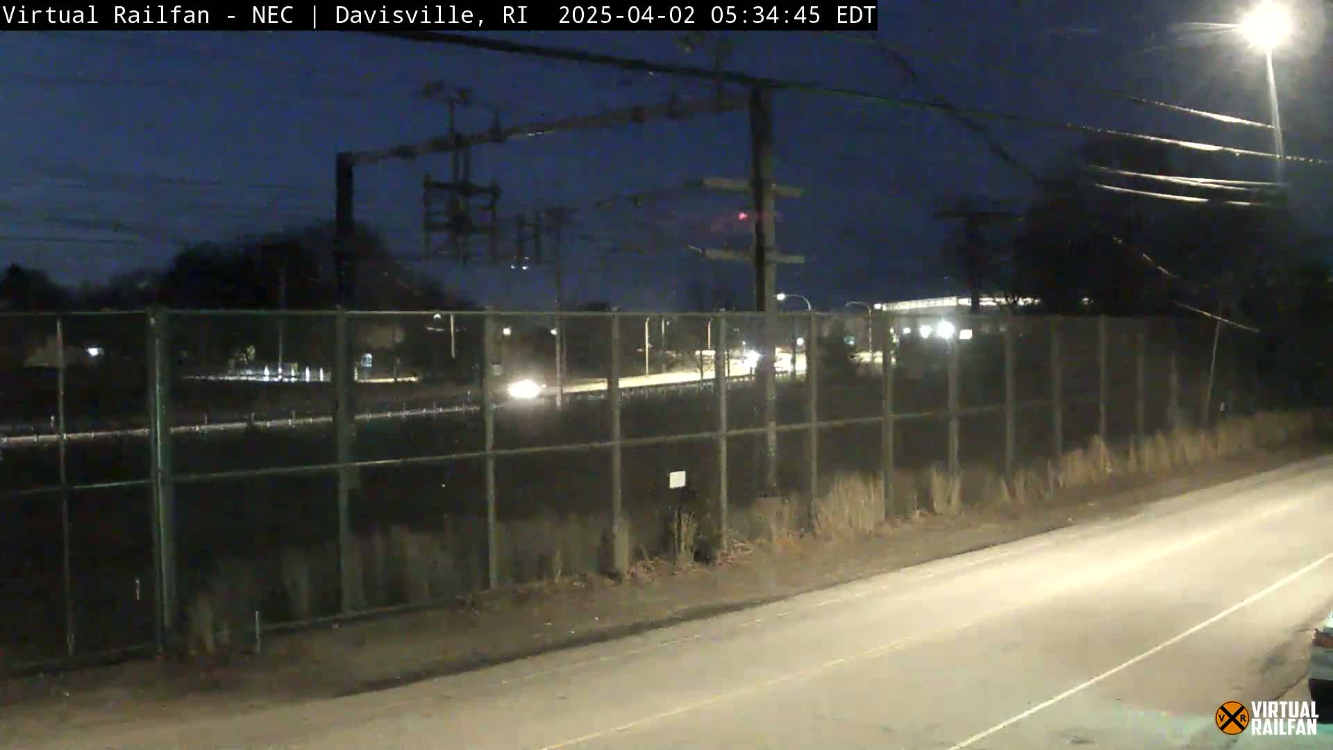 A nighttime view shows a road beside a chain-link fence separating it from a railway track, with a few cars visible in the distance and minimal lighting.