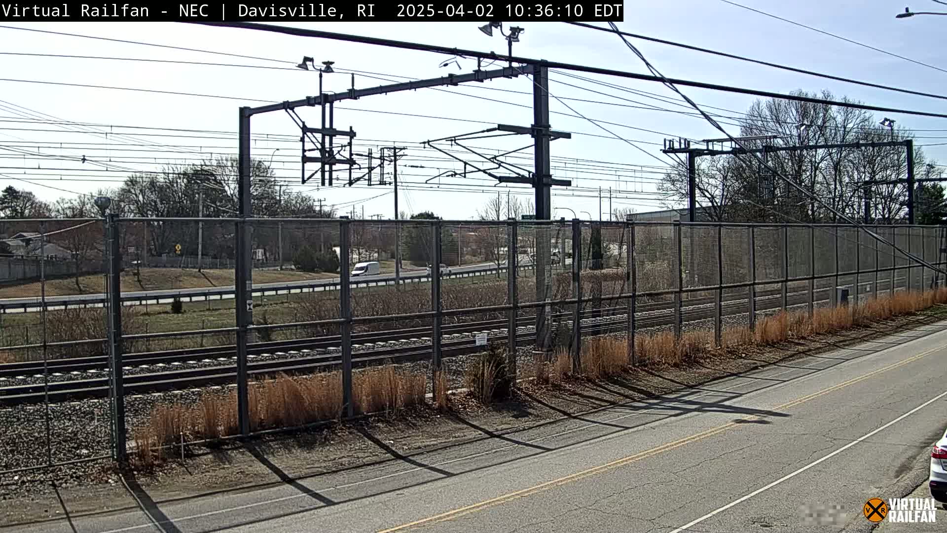 A view of railroad tracks beyond a chain-link fence, beside a road on a sunny day with bare trees and brown grasses.