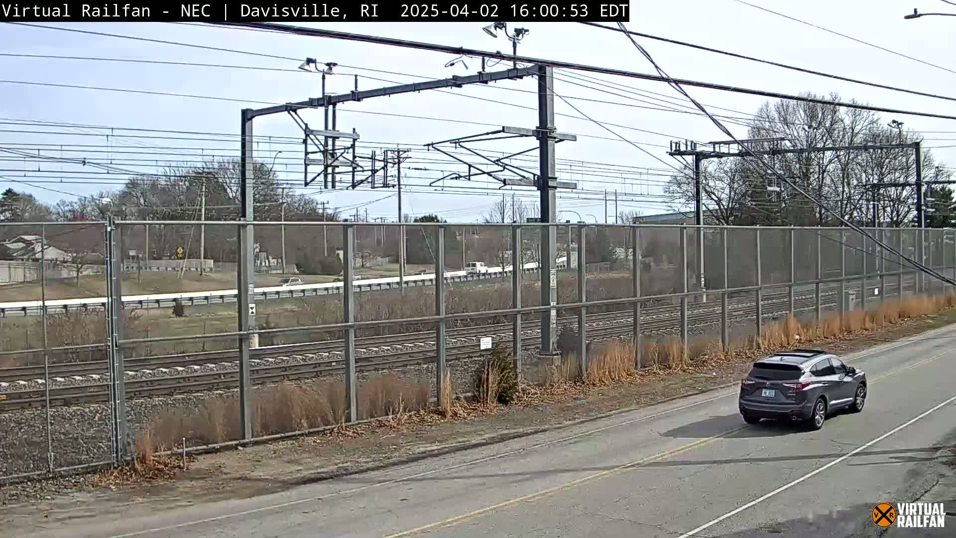 A gray SUV drives on a road next to a chain-link fence bordering a railway under a mostly clear sky.
