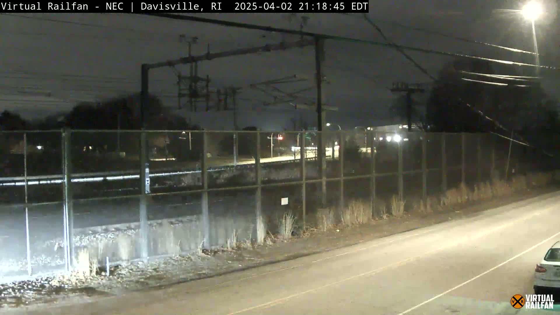 A nighttime view of a road next to a chain-link fence separating it from a railway, with sparse vegetation and some distant lights visible.