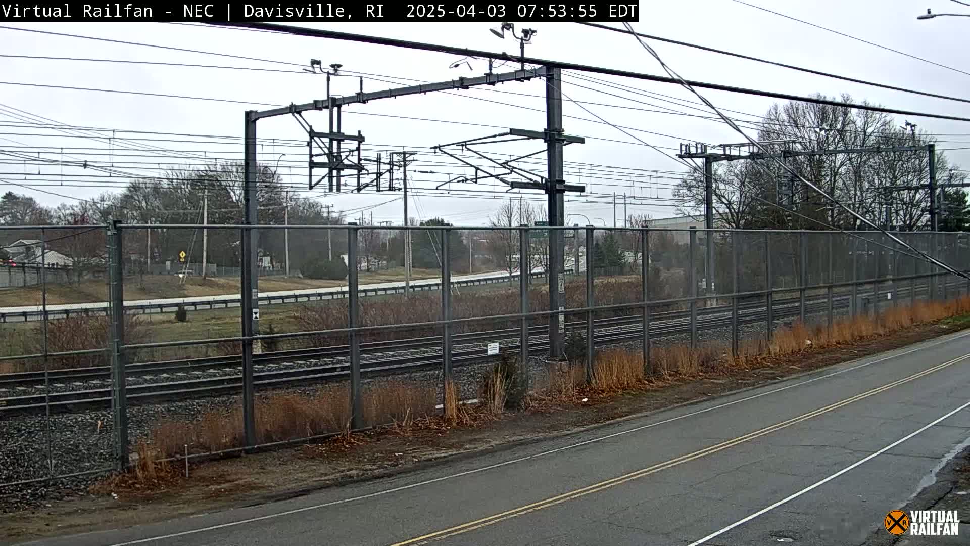 A view of railroad tracks behind a chain-link fence on an overcast day, with a road in the foreground.