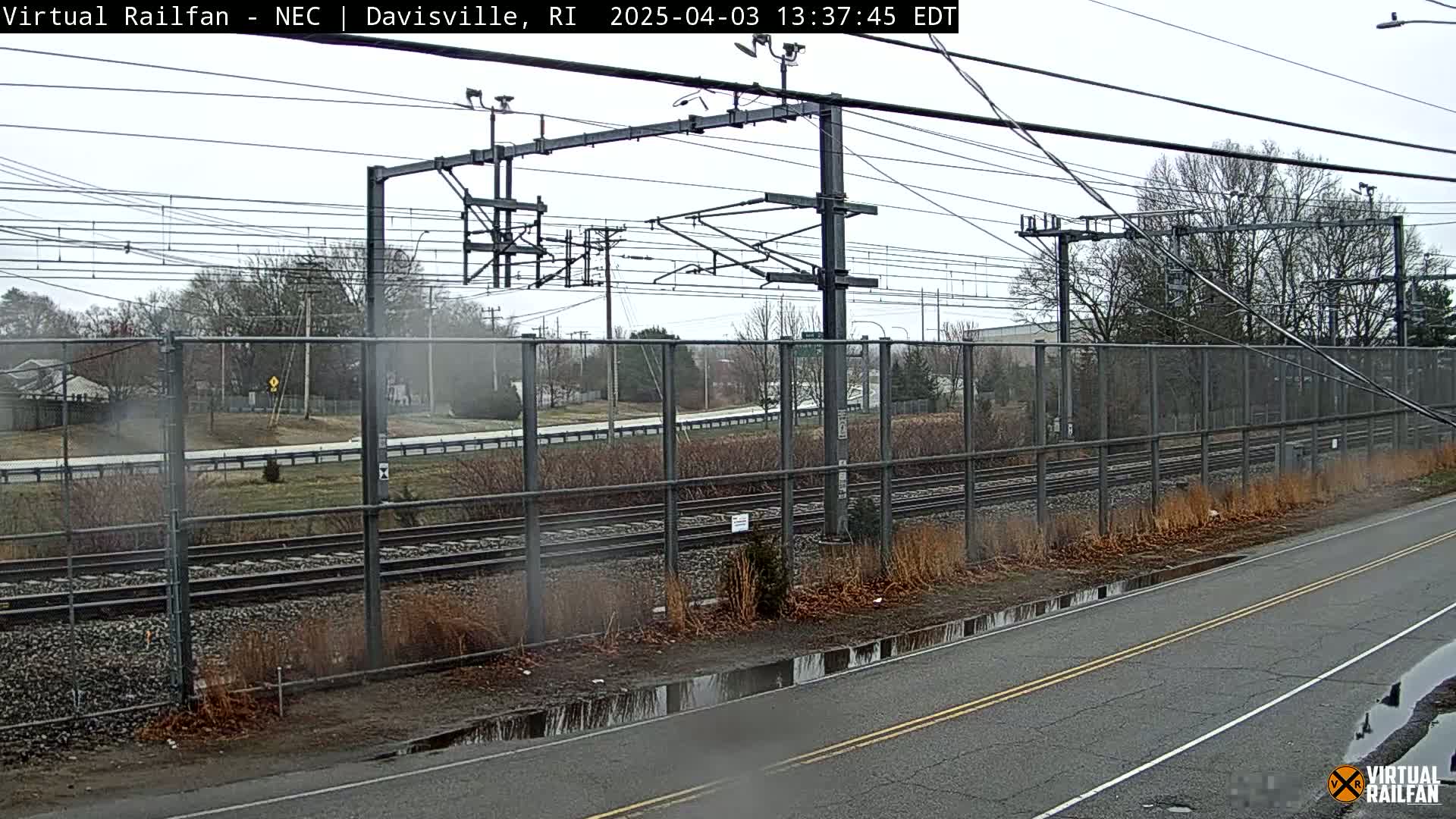 A view of a railway line behind a chain-link fence on an overcast day, with a road running alongside, showing puddles from recent rain.