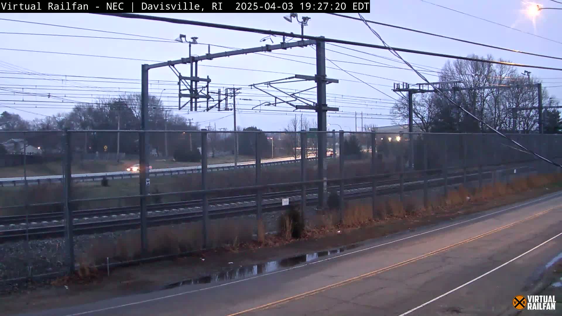 A view of train tracks next to a road under a twilight sky, with some puddles on the road and a chain link fence separating the tracks from the road.