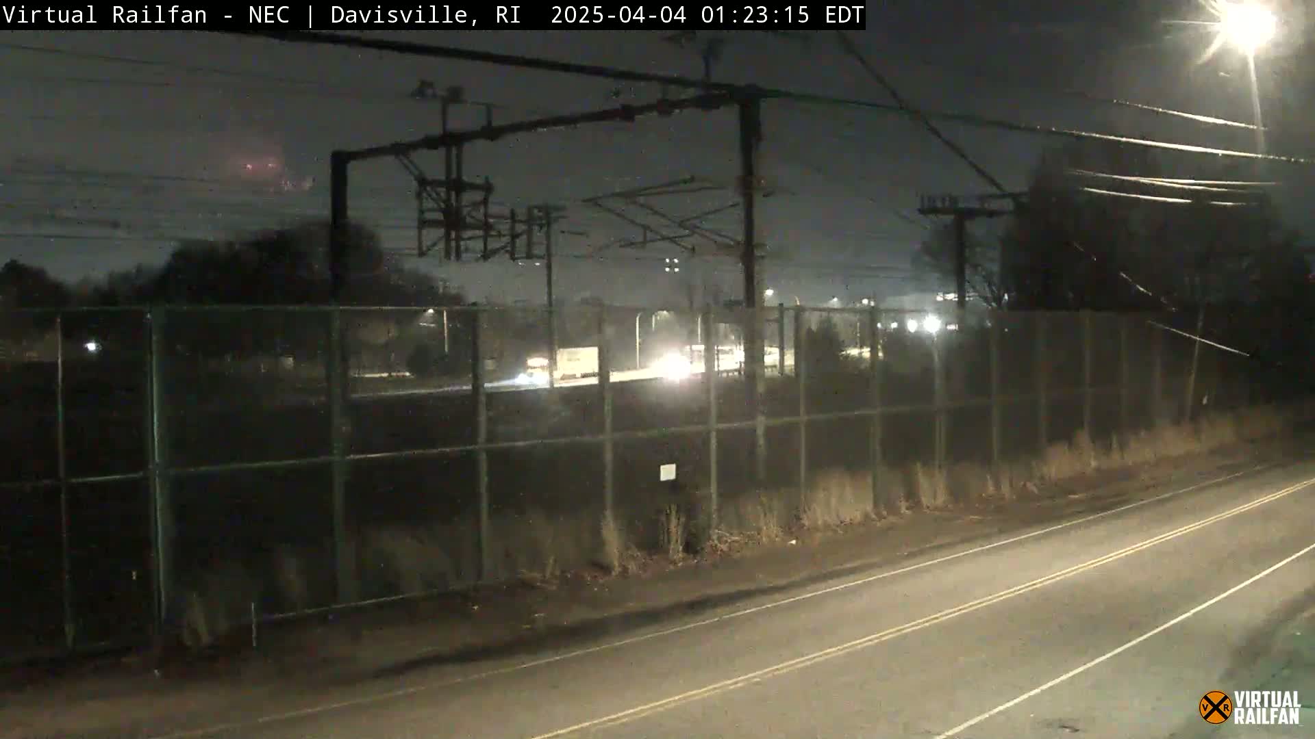 A nighttime view of a road and railway, beyond a chain-link fence, with vehicles visible in the distance under a dark sky.