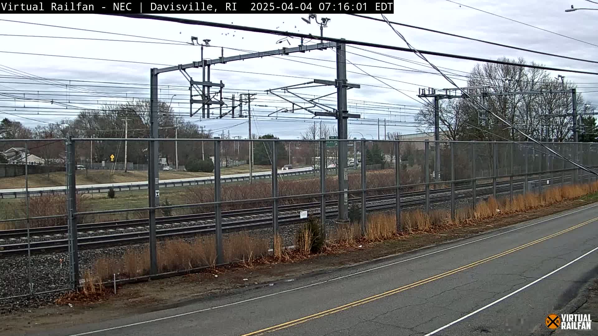 A view of train tracks behind a chain-link fence beside a road, under an overcast sky.