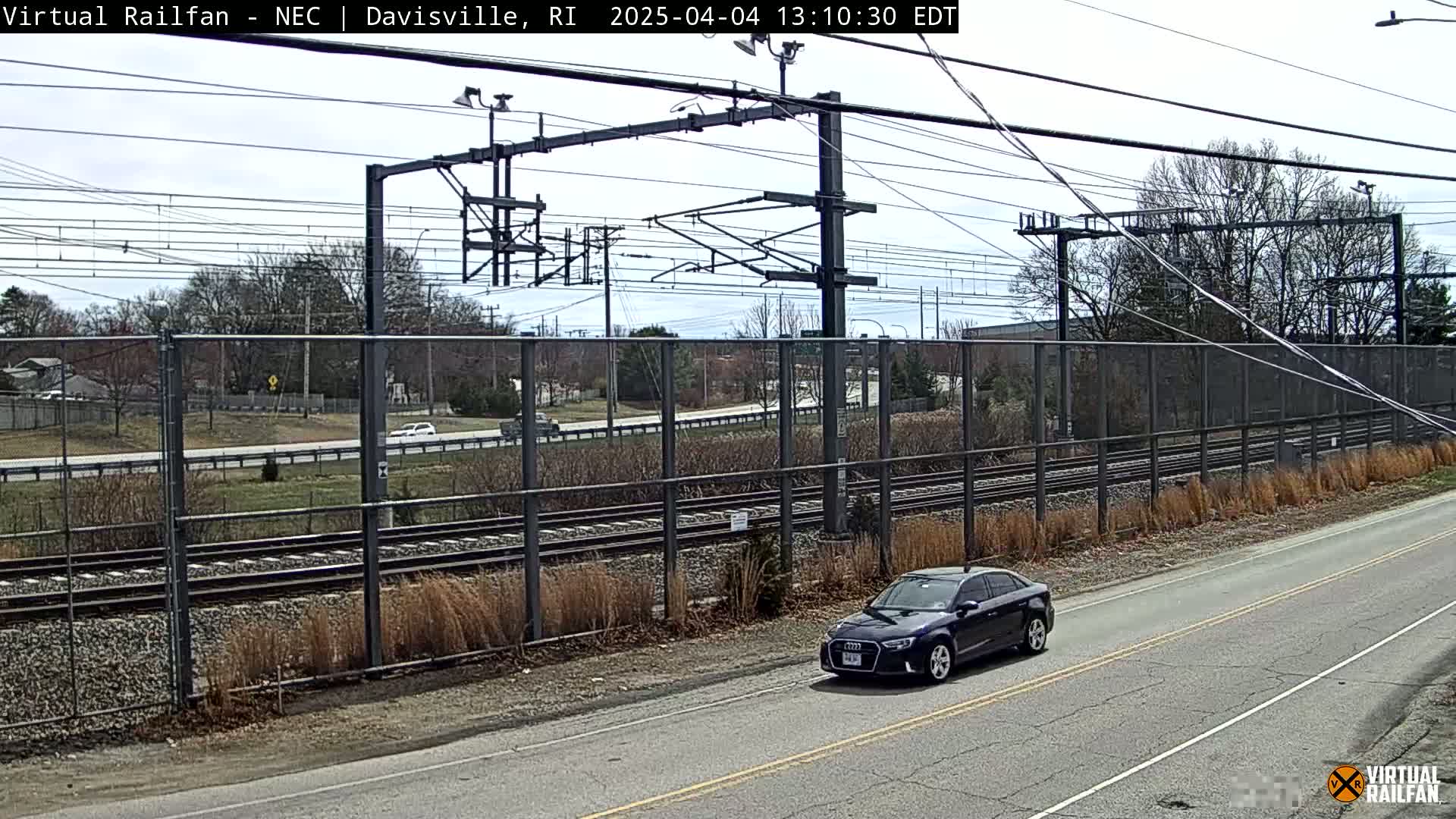 A dark-colored sedan drives on a road next to a railway under a partly cloudy sky.