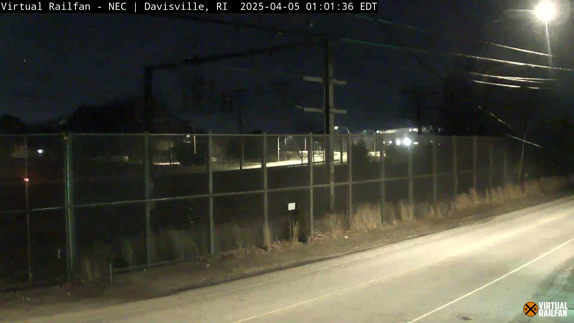 A nighttime view of a road next to a chain-link fence enclosing a dark area beyond it, with power lines overhead.