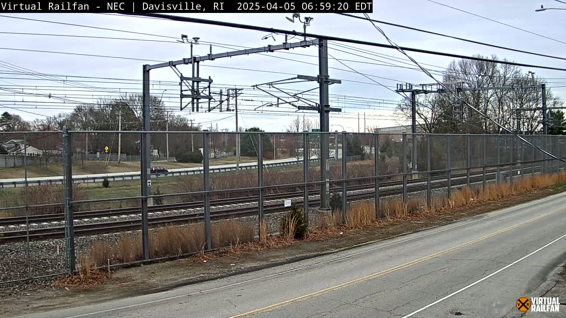 A view of train tracks behind a chain-link fence next to a road on an overcast day.