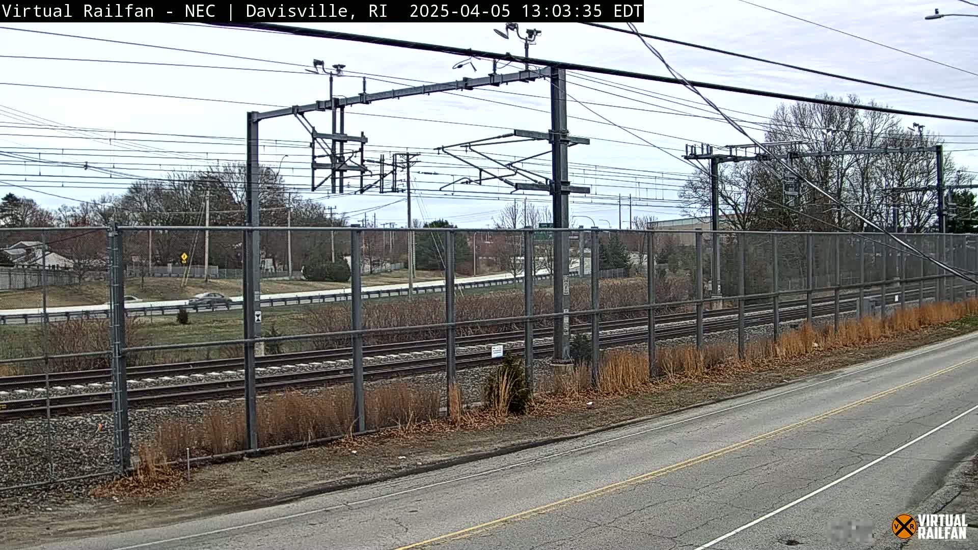 A view of railroad tracks behind a chain link fence beside a road on an overcast day.