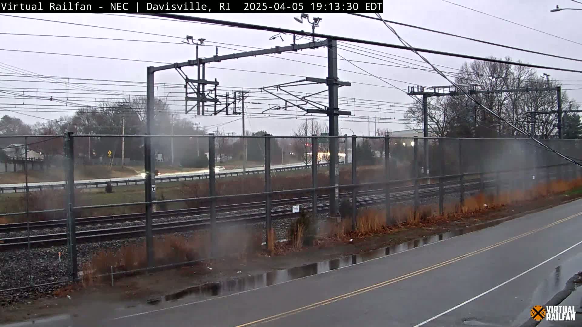 A wet road next to railroad tracks runs beside a chain-link fence separating it from a highway under an overcast sky.