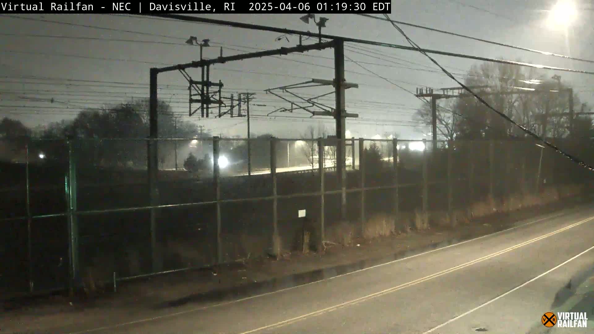 A nighttime view of a highway and train tracks separated by a chain-link fence, under rainy conditions.
