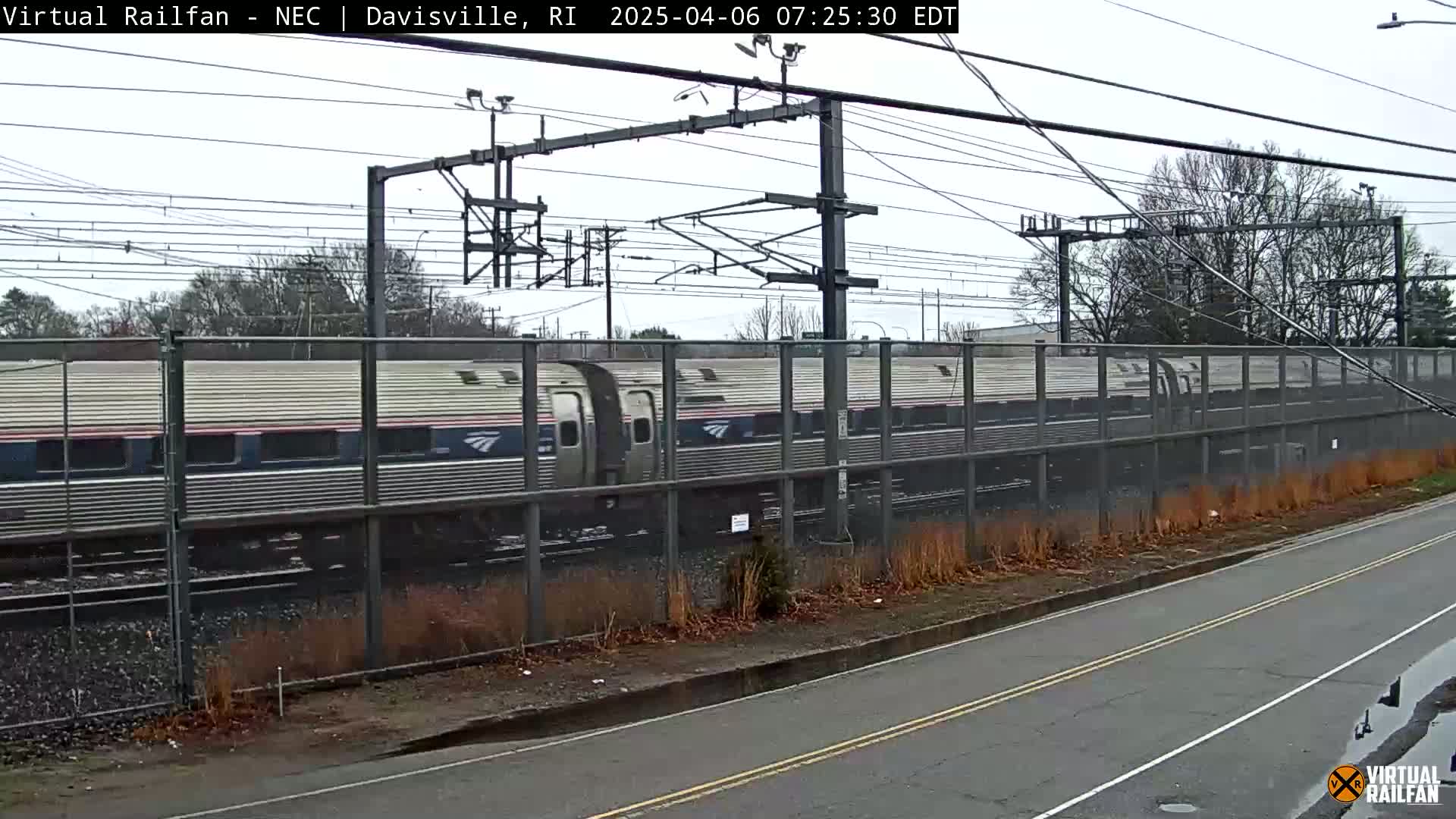 A long passenger train passes behind a chain-link fence on an overcast day.