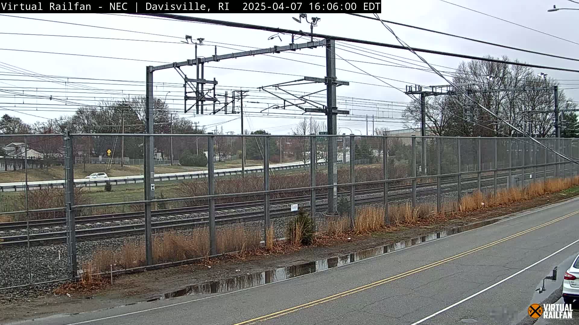 A view of train tracks behind a chain-link fence next to a road with puddles, under an overcast sky.