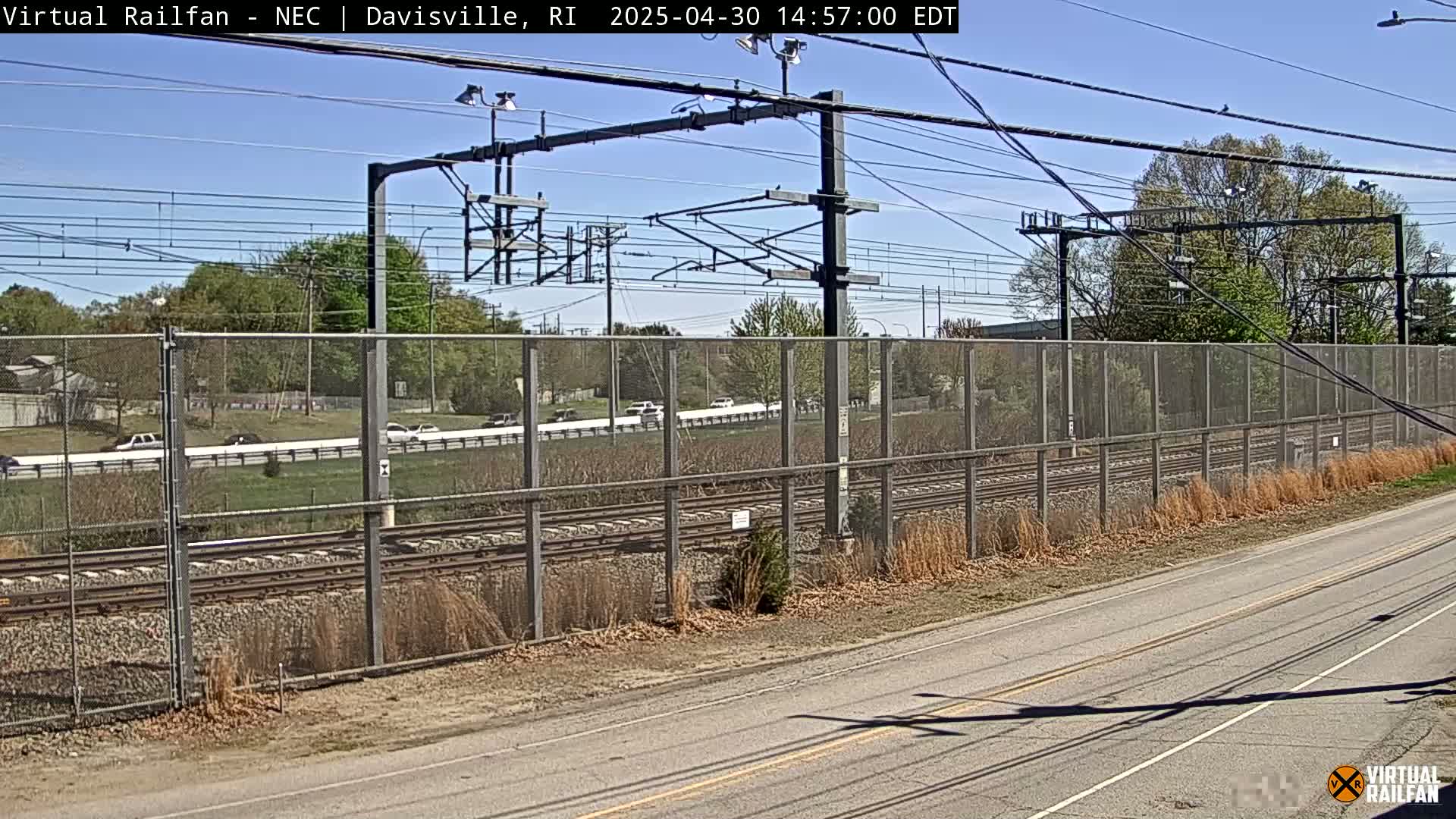 A sunny day reveals a view of railroad tracks behind a chain-link fence alongside a road, with cars visible on a road beyond the fence and trees lining the background.