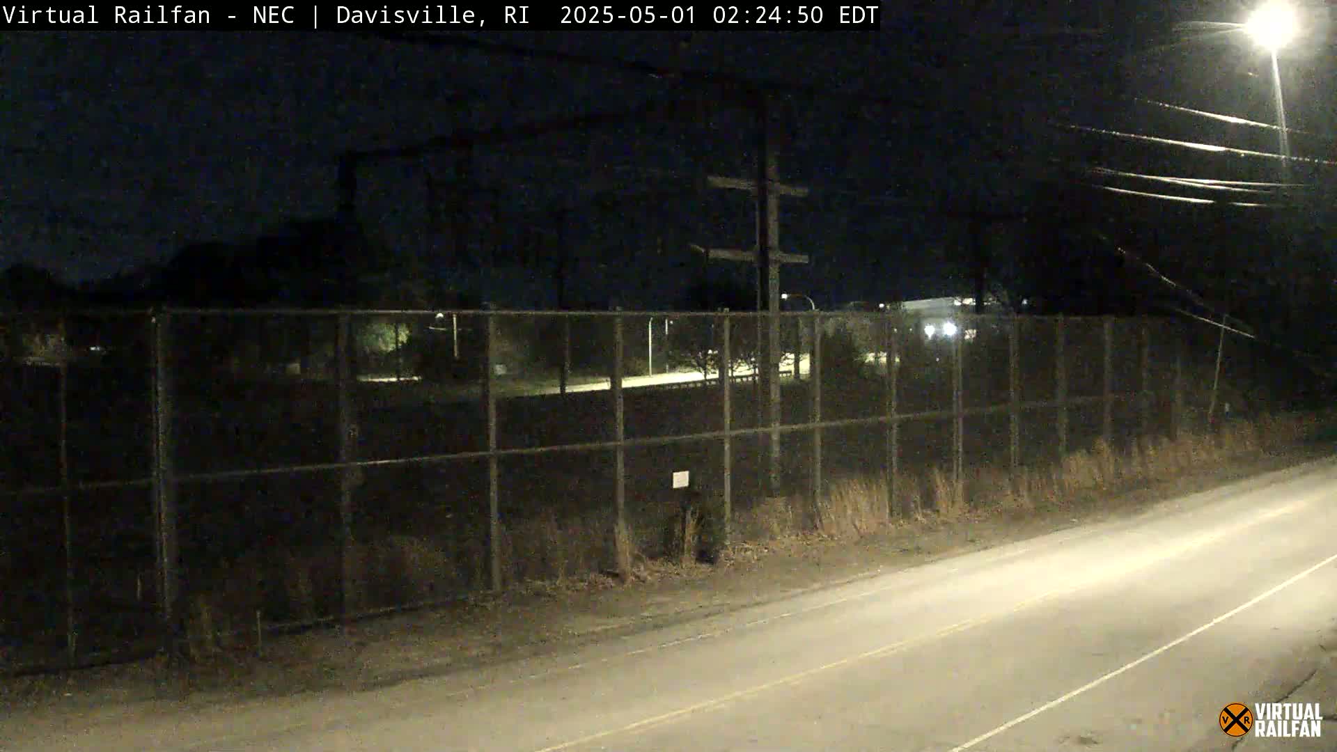 A nighttime view of a chain-link fence enclosing a dark area, beside a road lit by a streetlight.