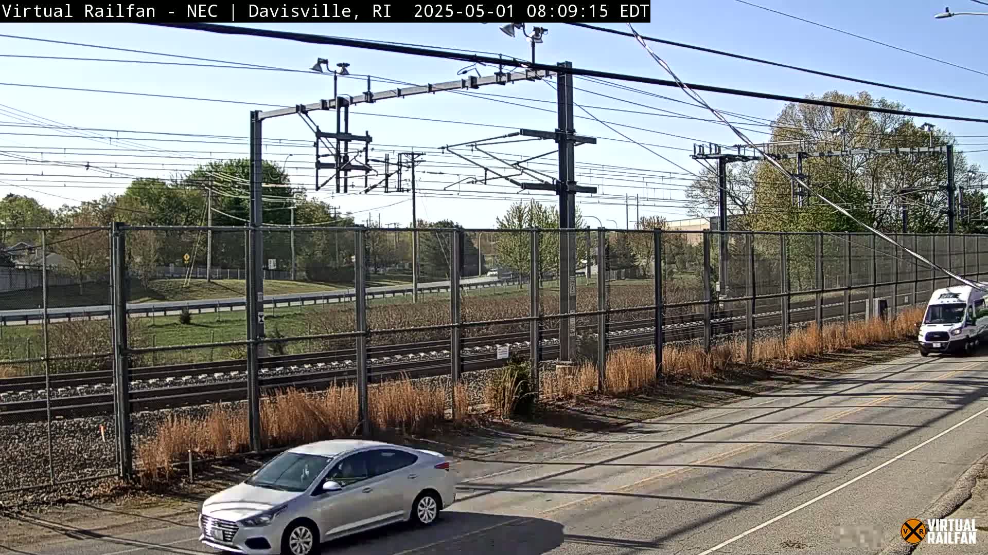 A silver car and a white van are parked on a road next to a chain-link fence enclosing railroad tracks under a sunny sky.