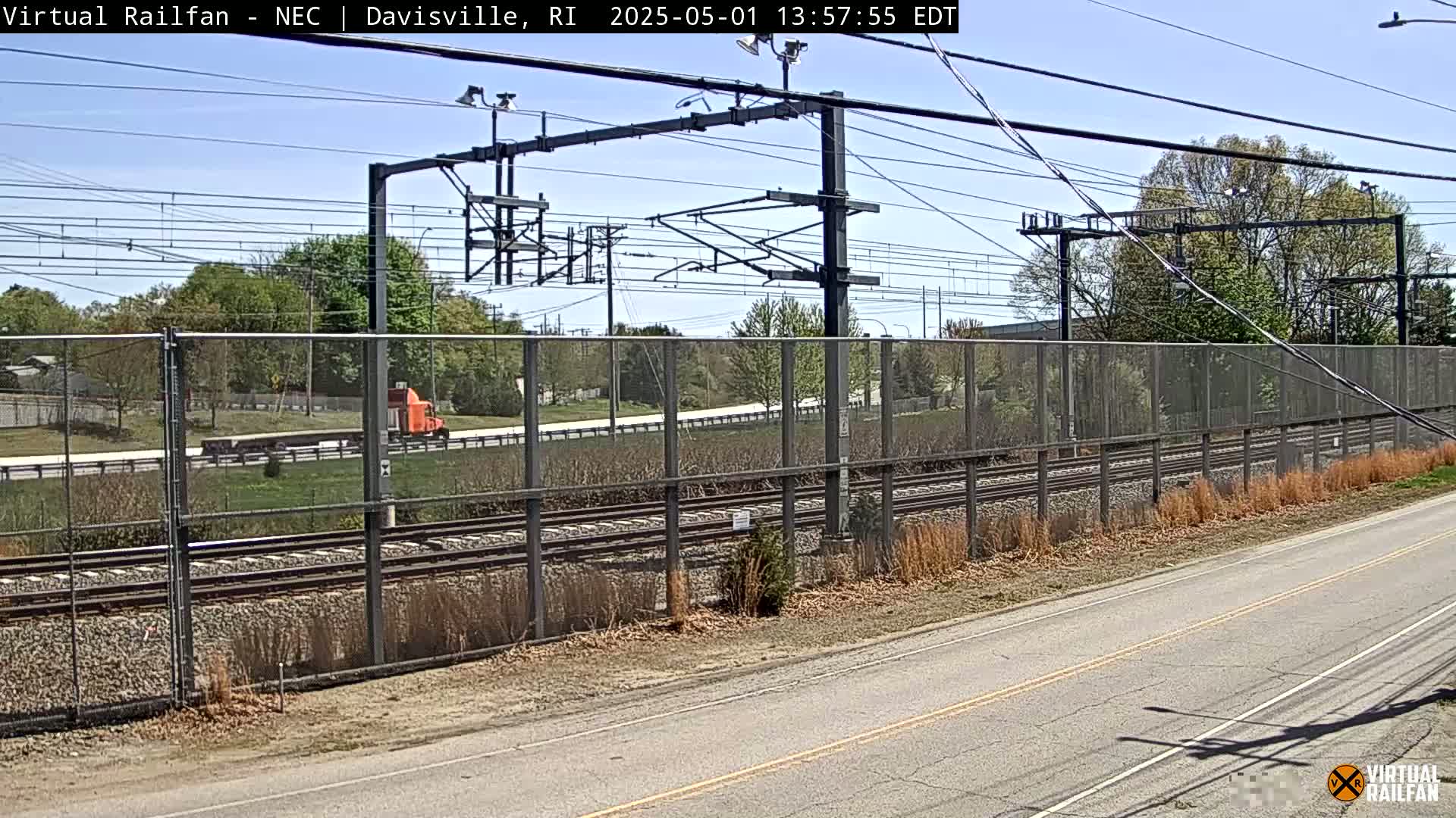 A semi-truck drives past a section of railway tracks on a sunny day, beyond a chain-link fence.