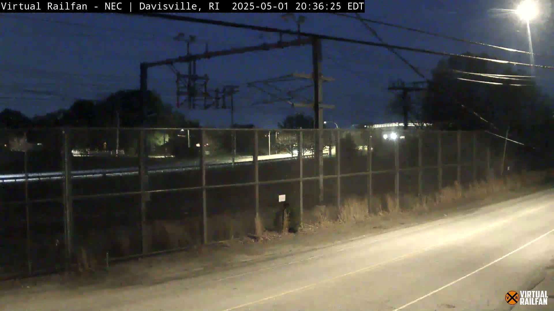 A nighttime view shows a chain-link fence separating a road from a railway, with overhead electrical lines and sparse vegetation.
