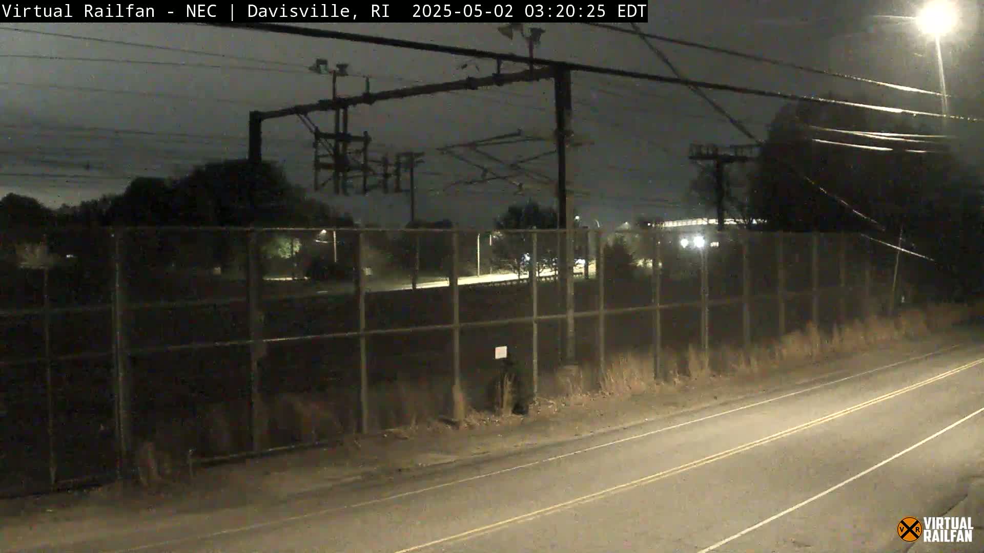 A nighttime view shows a road next to a chain-link fence enclosing a rail yard under a dark, mostly clear sky.