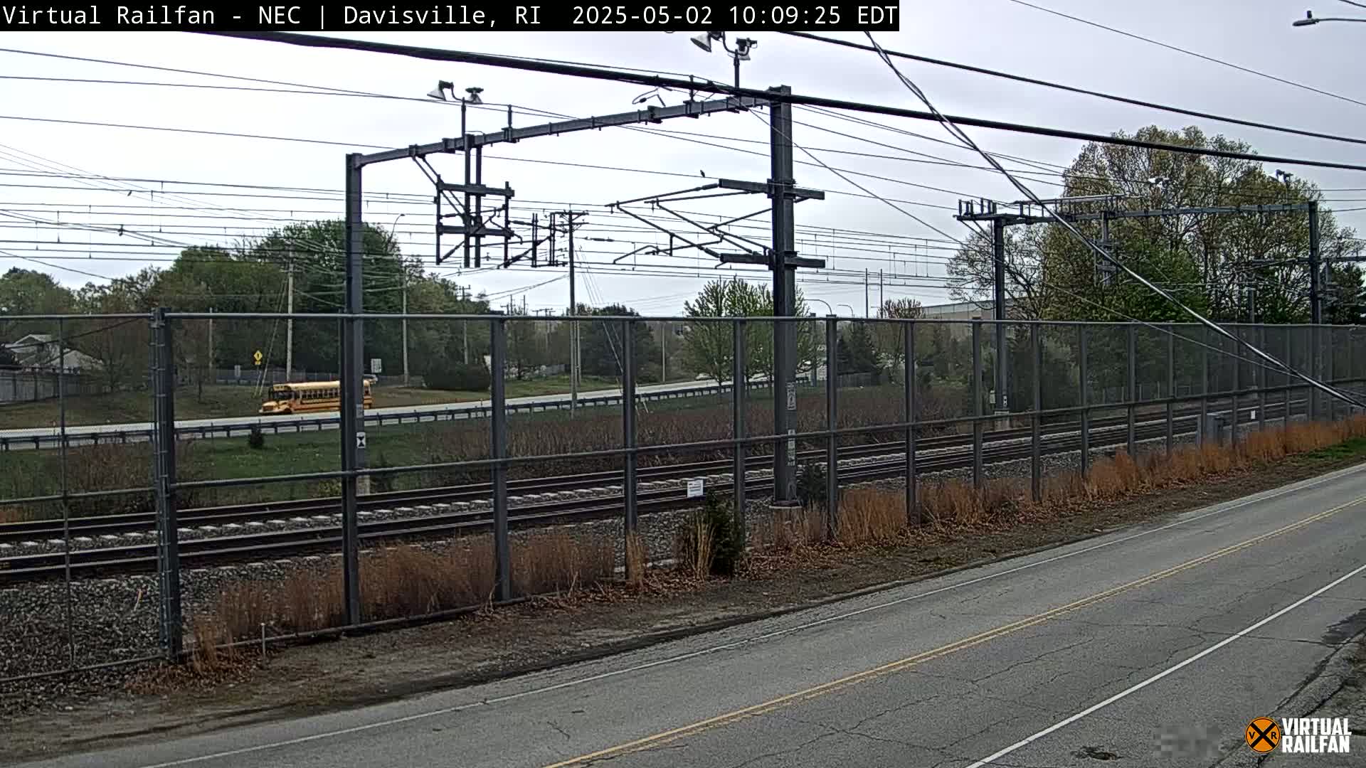 A view of railroad tracks behind a chain-link fence, next to a road with a school bus visible in the distance, under an overcast sky.