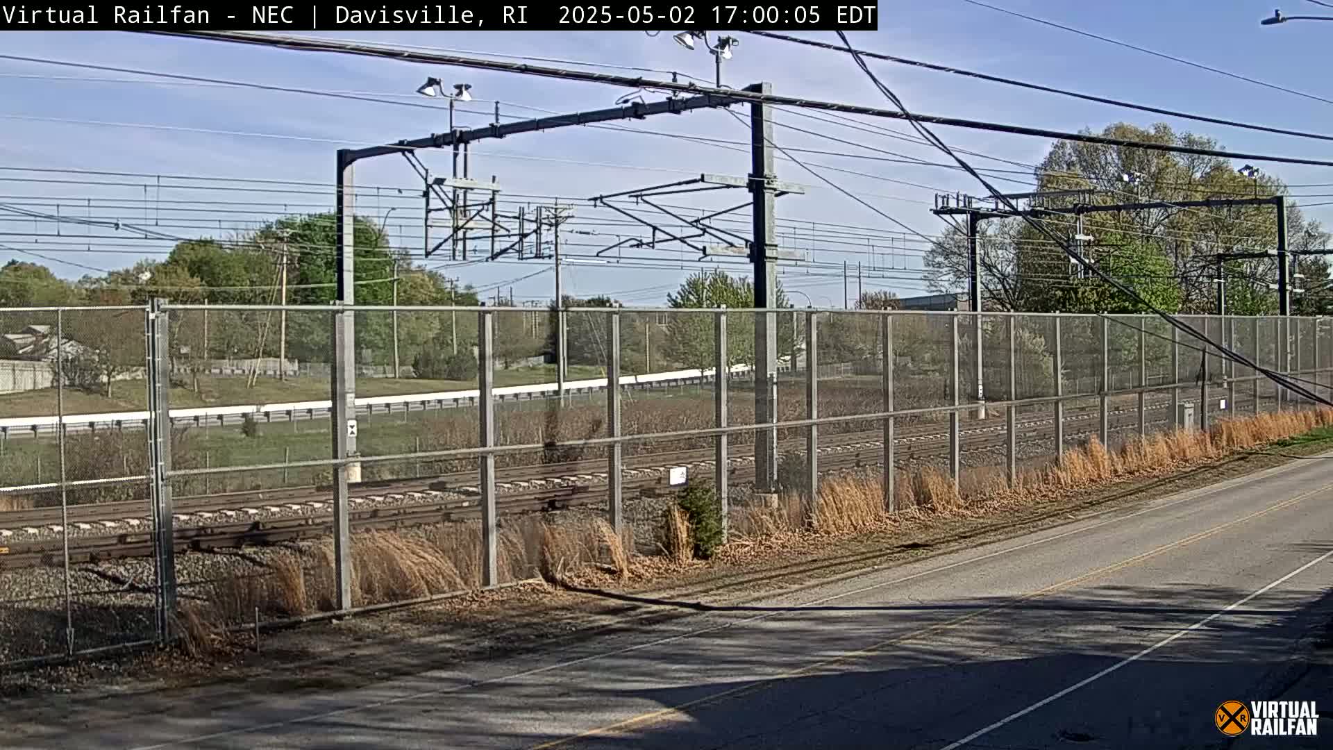 A view of railroad tracks behind a chain link fence on a sunny day, with trees and a road in the foreground.