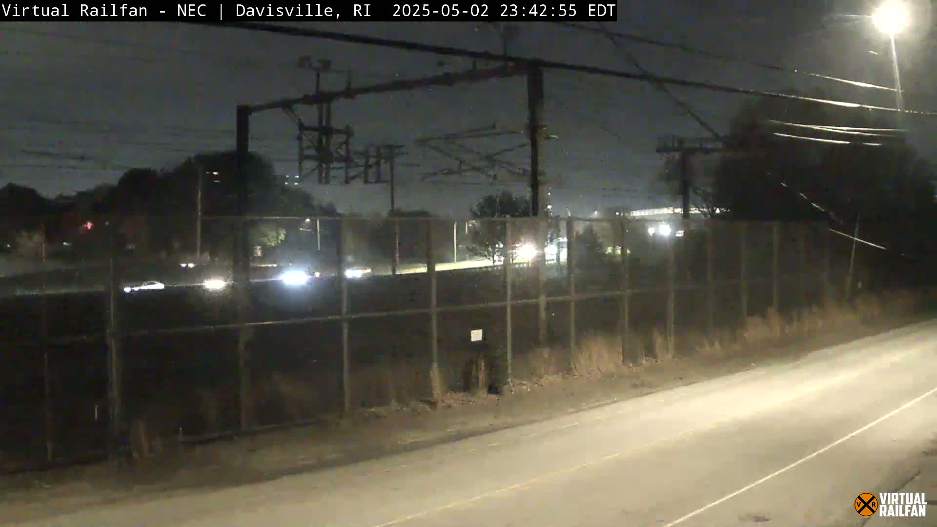 A nighttime view of a road next to a chain-link fence, with sparse traffic visible beyond the fence under a dark sky.
