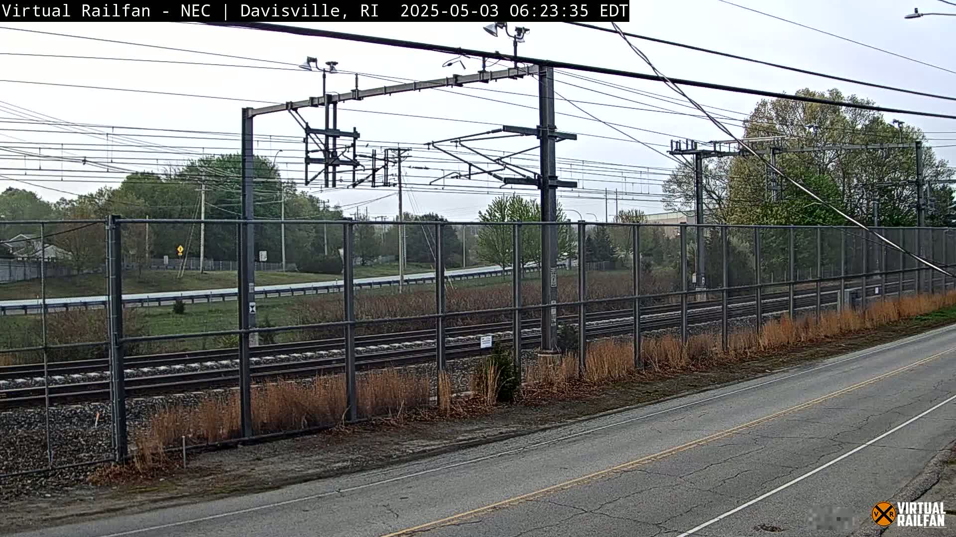 A section of railway tracks runs behind a chain link fence, next to a road, under an overcast sky.
